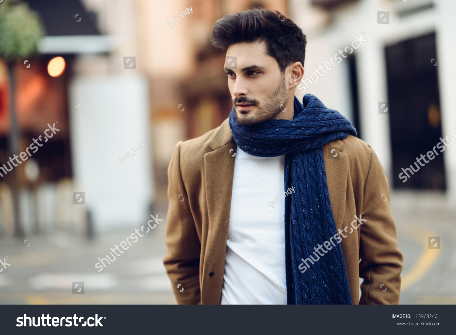 Young man wearing winter clothes in the street. Young bearded guy with modern hairstyle with coat, scarf, blue jeans and t-shirt. #1139682401