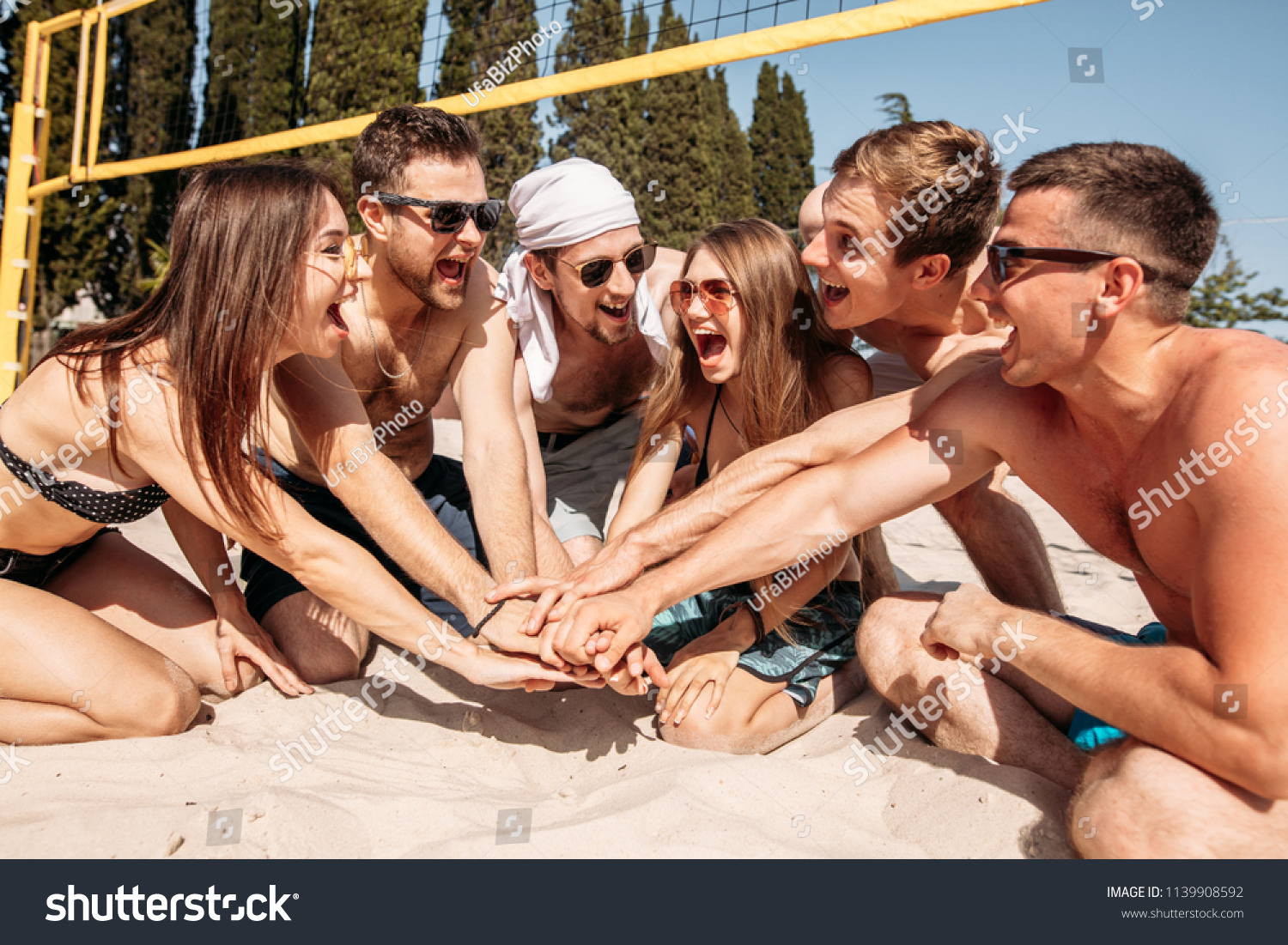 Give high five. Group of fit excited caucasian people talking in a circle, sitting on sand after volleyball game won. discussing goal, achieving team results. Teamwork, vacation, active life benefit #1139908592