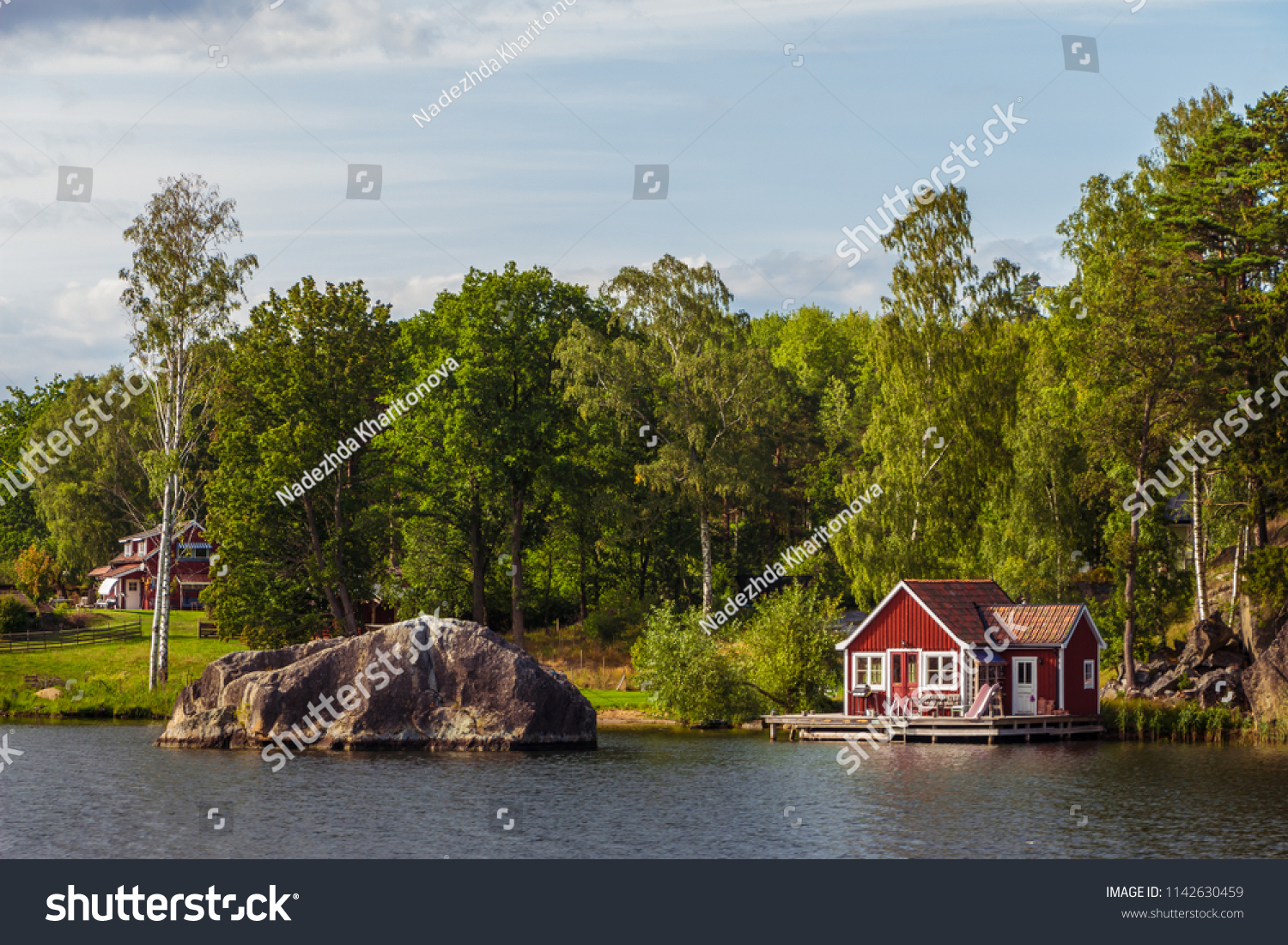 View on red holiday cabin by a lake in Stockholm archipelago, Sweden. Wooden cottage, sauna on shore. Tiny house near the water. Rocky small island, islet in water. Buildings surrounded by green trees #1142630459