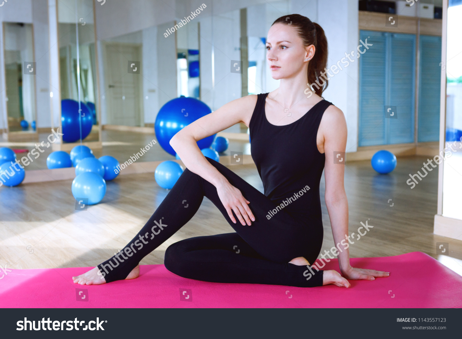 Woman sitting on pink ypga matt and doing exercises in fitness club gym, wearing black overall jumpsuit #1143557123