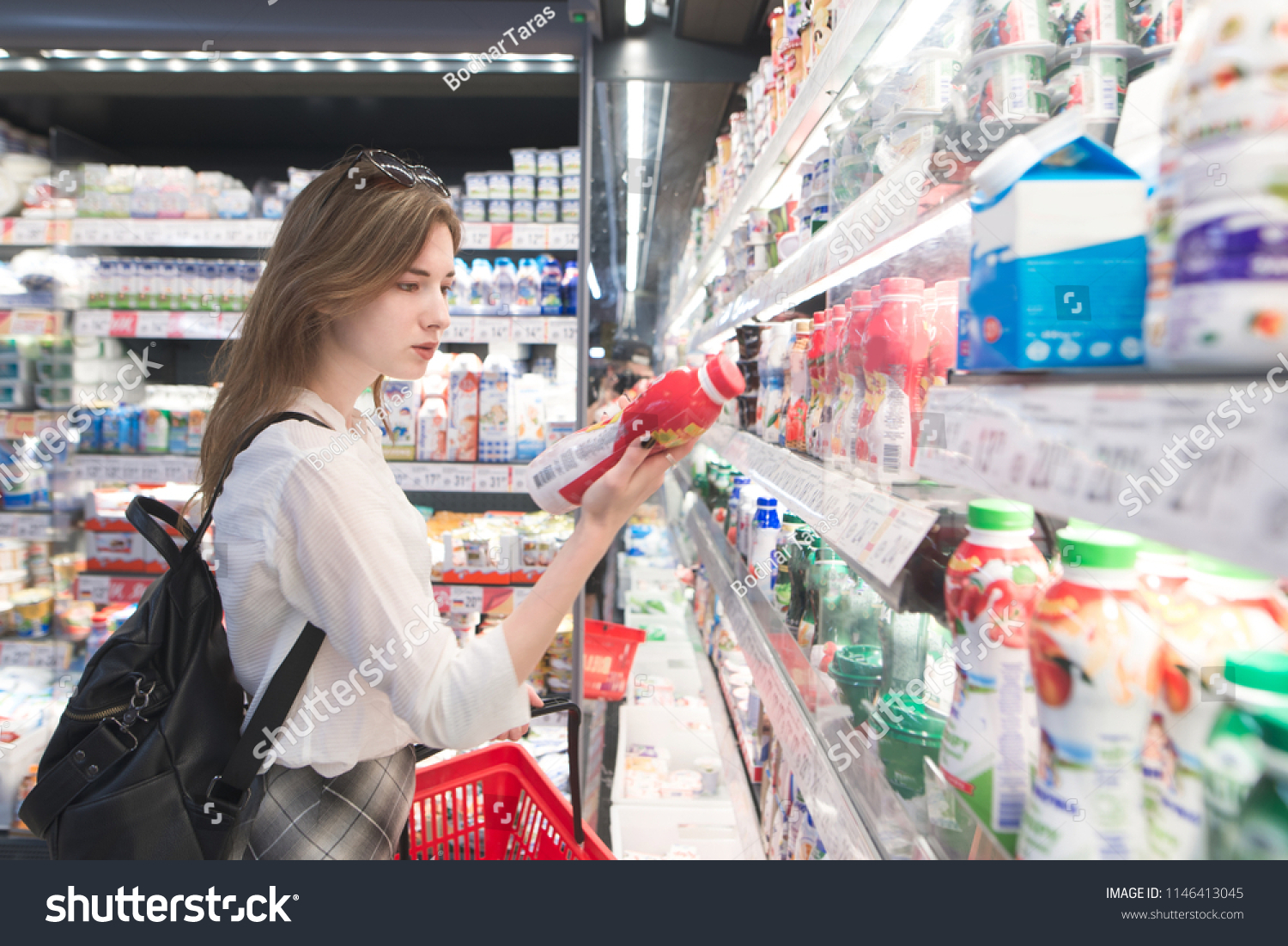 Stylish attractive woman is standing by the refrigerator with dairy products, holds yogurt in his hands and reads the label. An attractive girl buys a yogurt in a supermarket. #1146413045