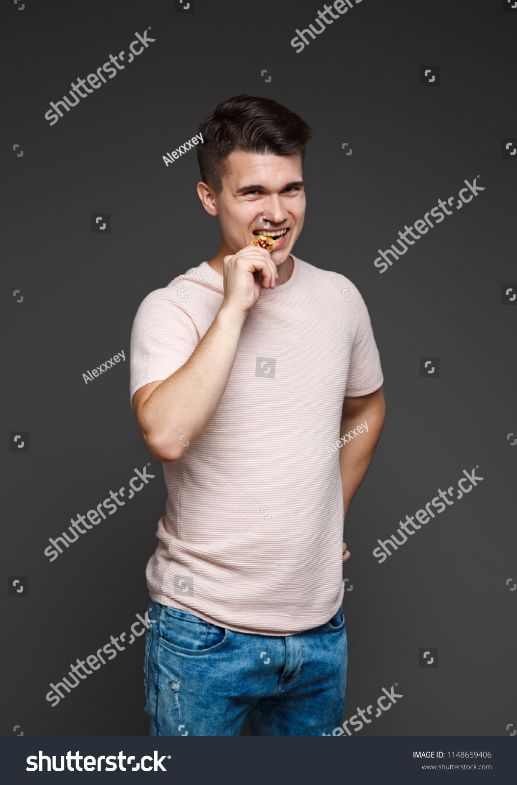 young guy bites a coin bitcoin with his teeth, checking for authenticity, isolated on a gray background. #1148659406
