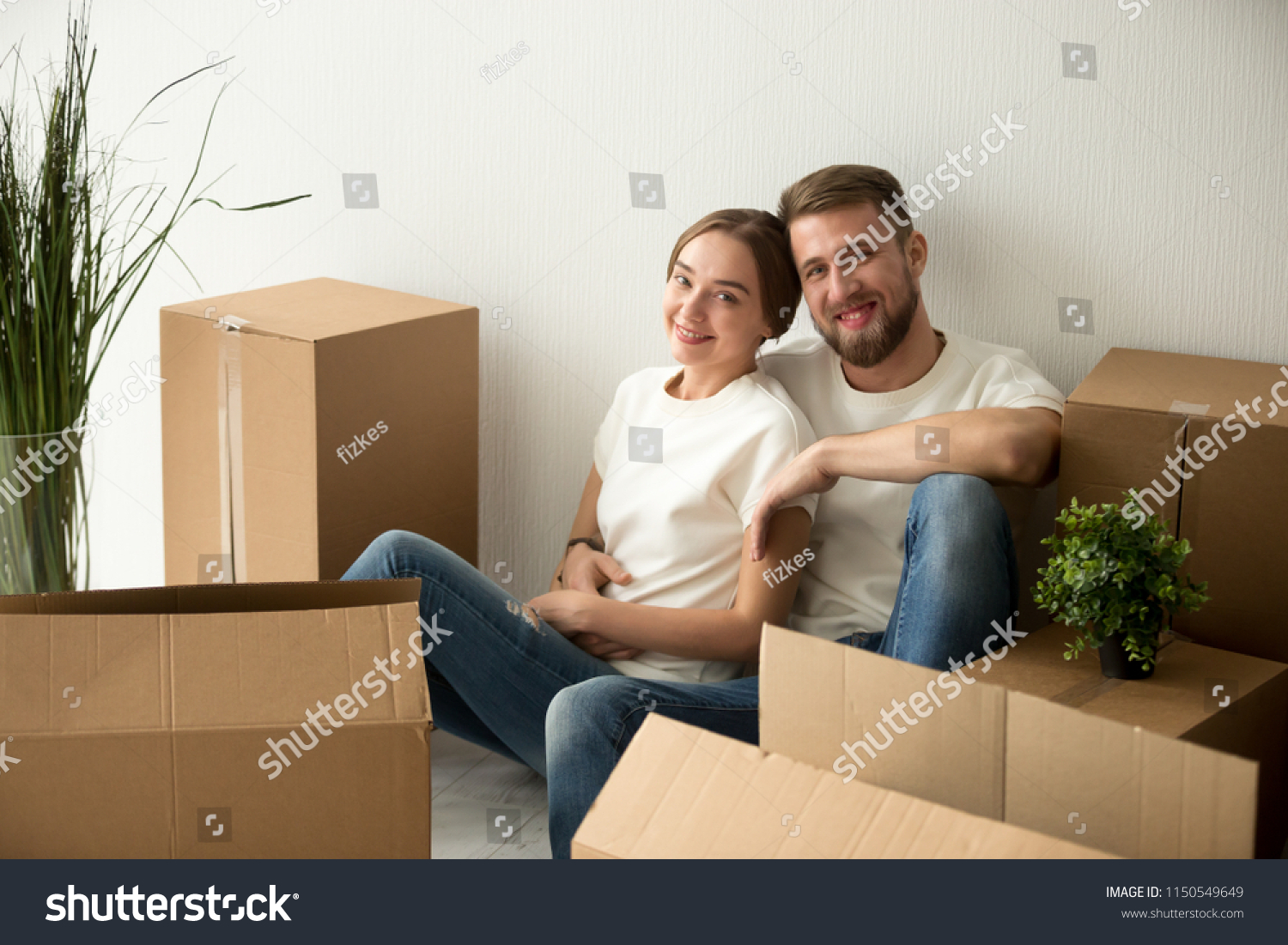 Portrait of smiling millennial spouses pose near cardboard boxes after moving in to first bought home, happy couple look at camera sitting on floor in new family house hugging, enjoying relocation day #1150549649