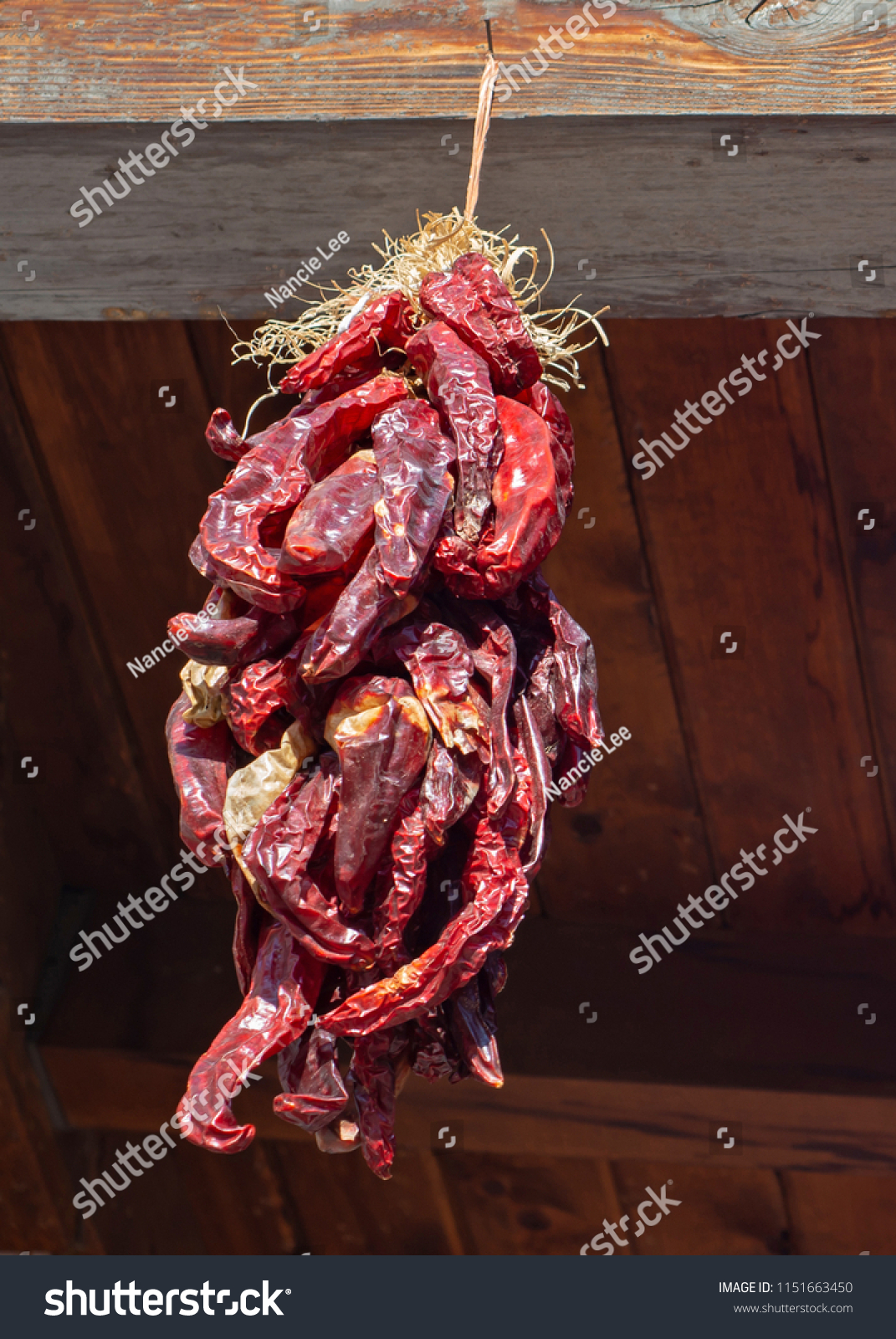 A long ristra of dried red chilis hanging from a beam outdoors.  #1151663450