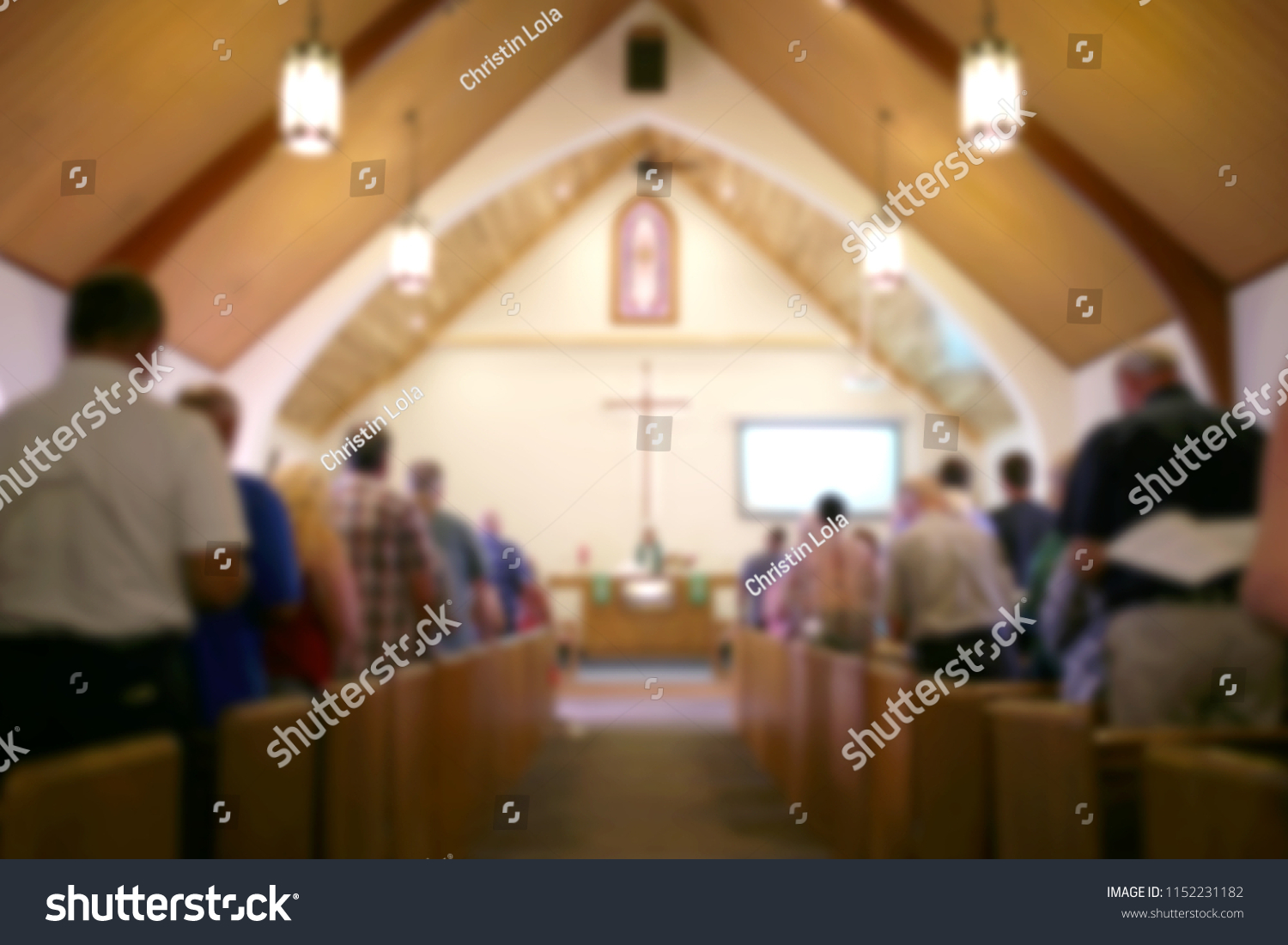 A blurred photo of the inside of a church sanctuary that is filled with people in the pews, and the pastor stands under a large cross at the altar. #1152231182