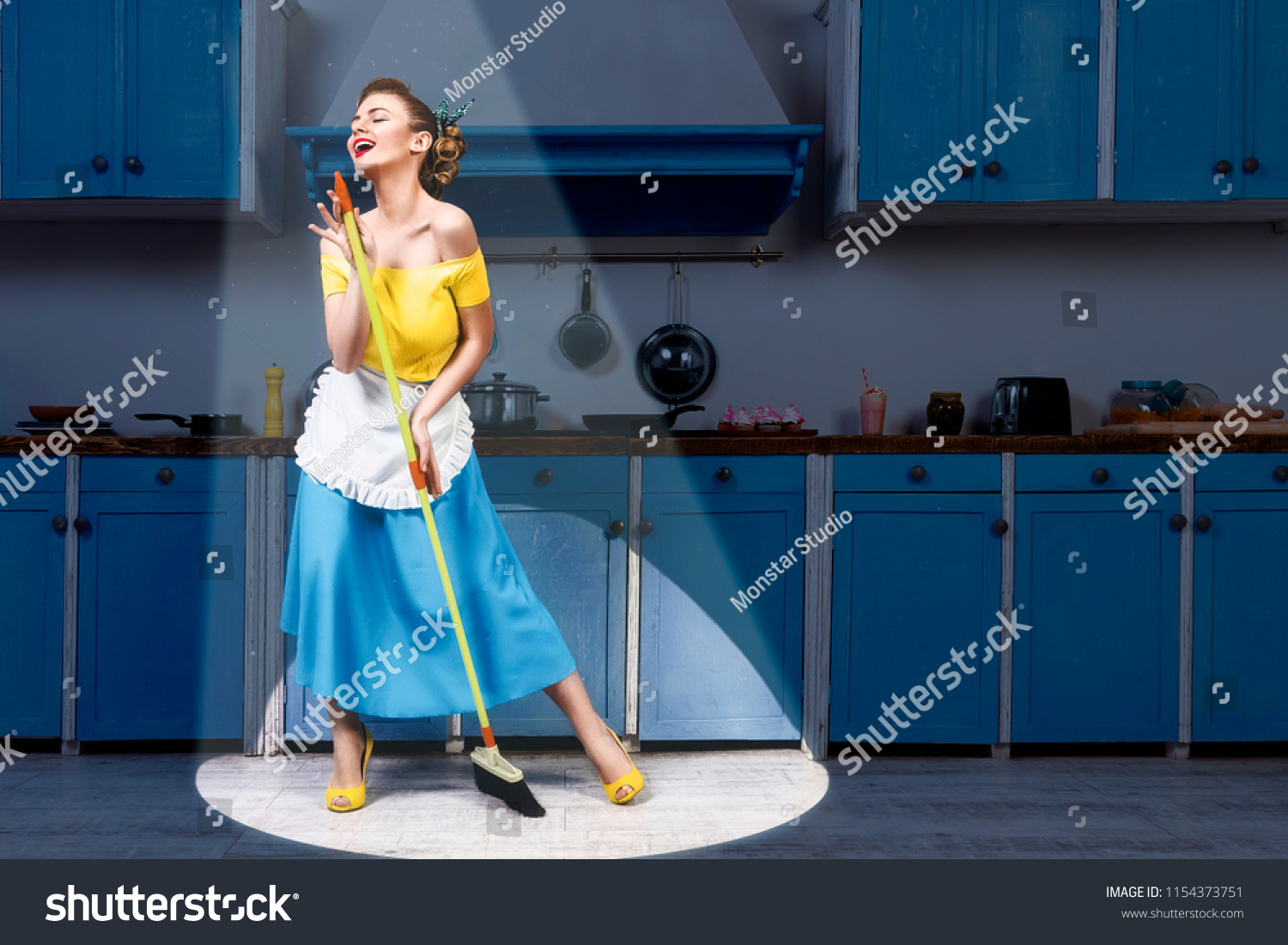 Retro pin up girl woman female housewife wearing yellow top, blue skirt and white apron holding mop singing and cleaning floor in stage light kitchen with blue cabinets and utensils. Housework concept #1154373751