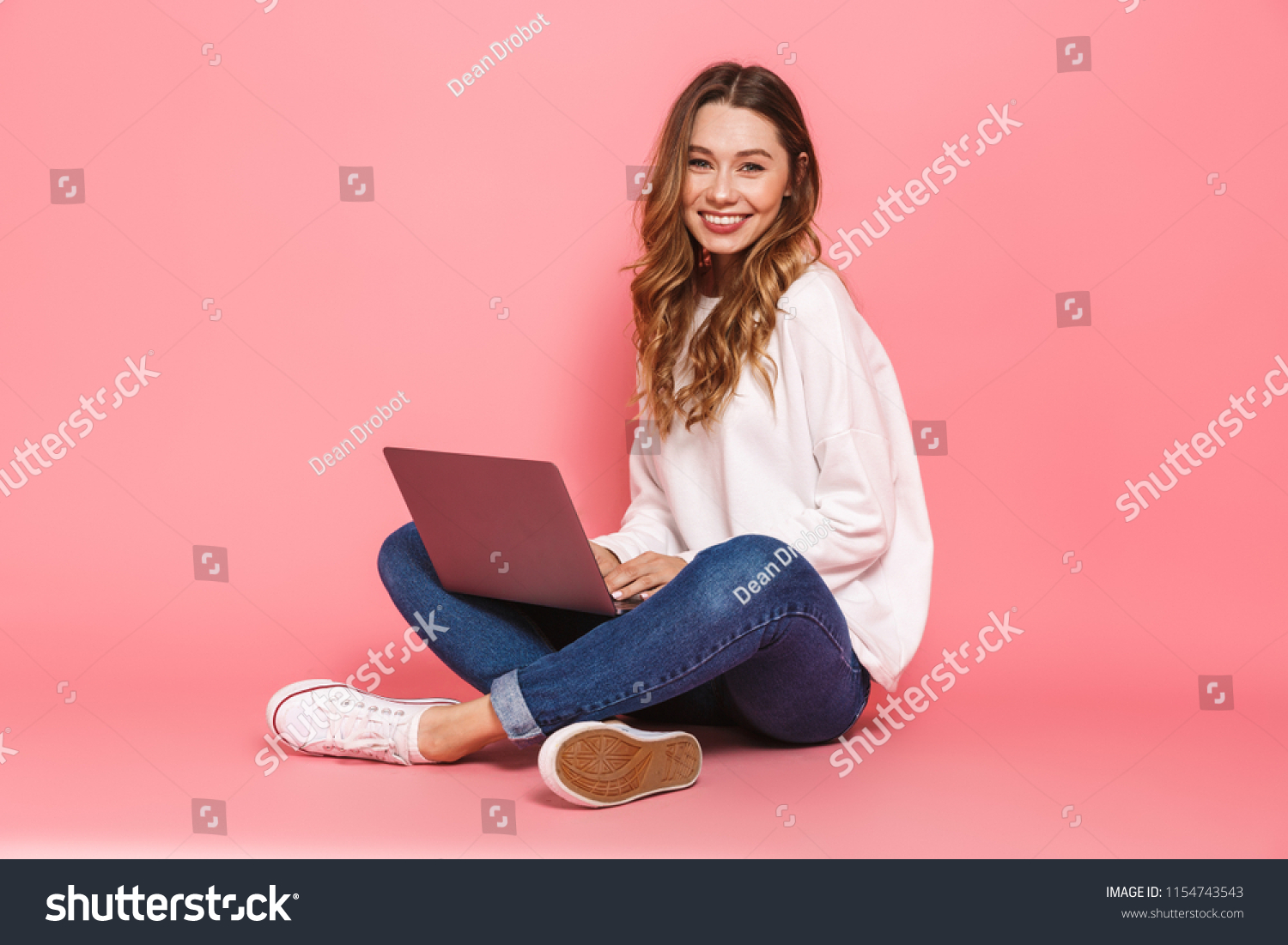 Portrait of a smiling young woman sitting with legs crossed, using laptop computer isolated over pink background #1154743543