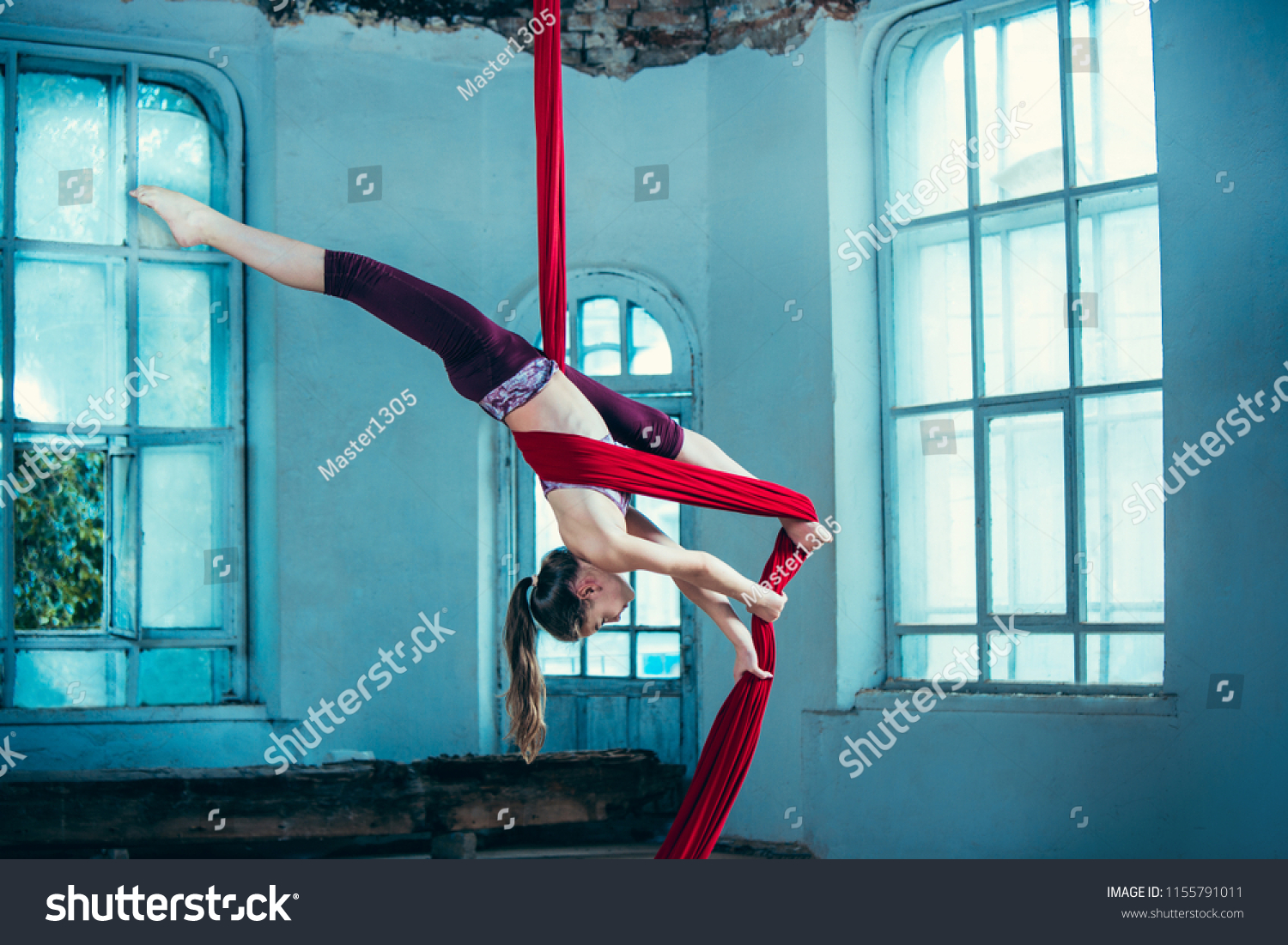 Graceful gymnast performing aerial exercise with red fabrics on blue old loft background. Young teen caucasian fit girl. The circus, acrobatic, acrobat, performer, sport, fitness, gymnastic concept #1155791011