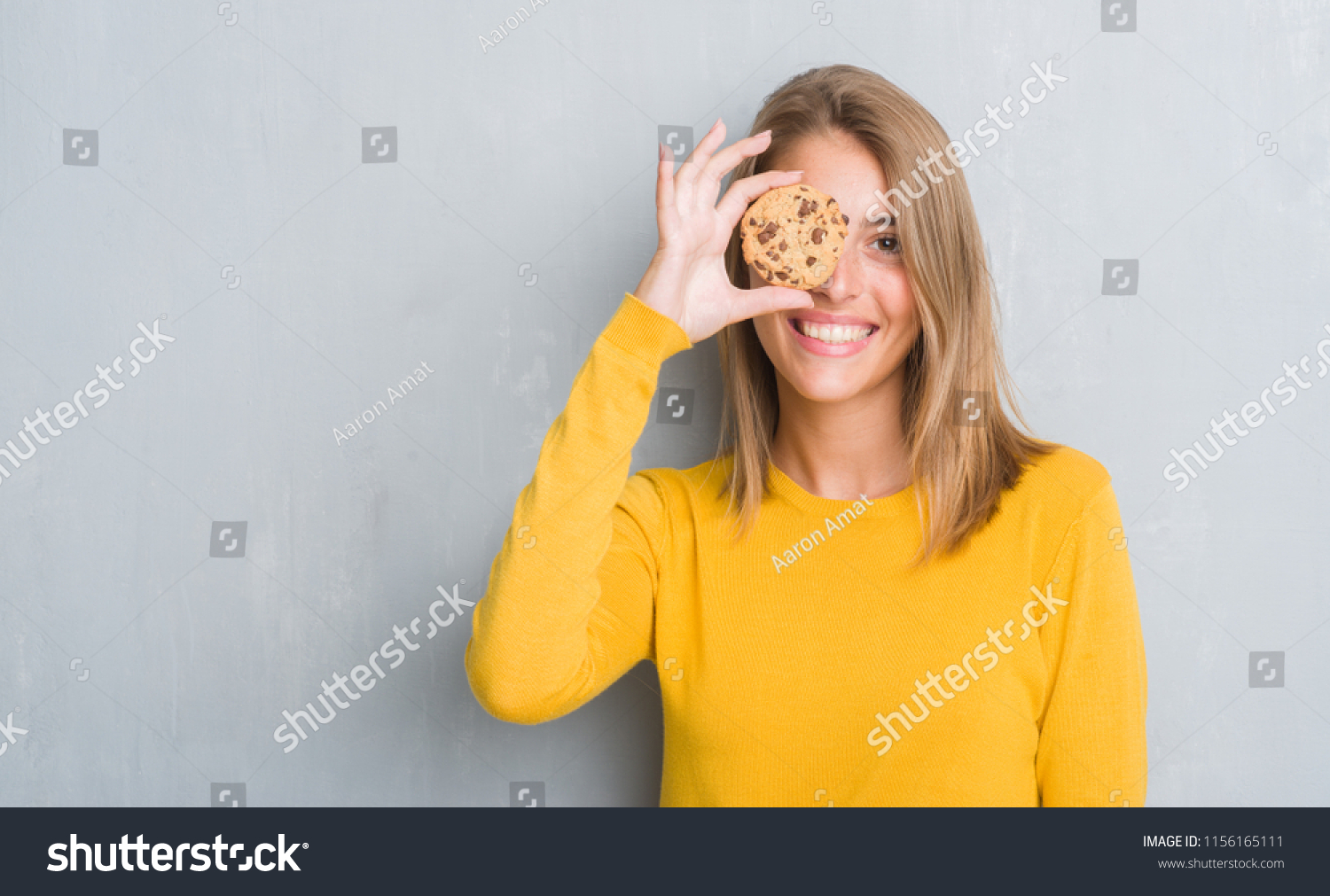 Beautiful young woman over grunge grey wall eating chocolate chip cooky with a happy face standing and smiling with a confident smile showing teeth #1156165111