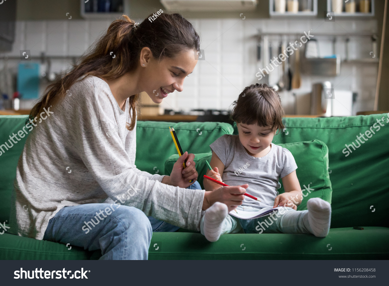 Smiling baby sitter and preschool kid girl drawing with colored pencils sitting on sofa together, single mother and child daughter playing having fun, creative family activities at home concept #1156208458