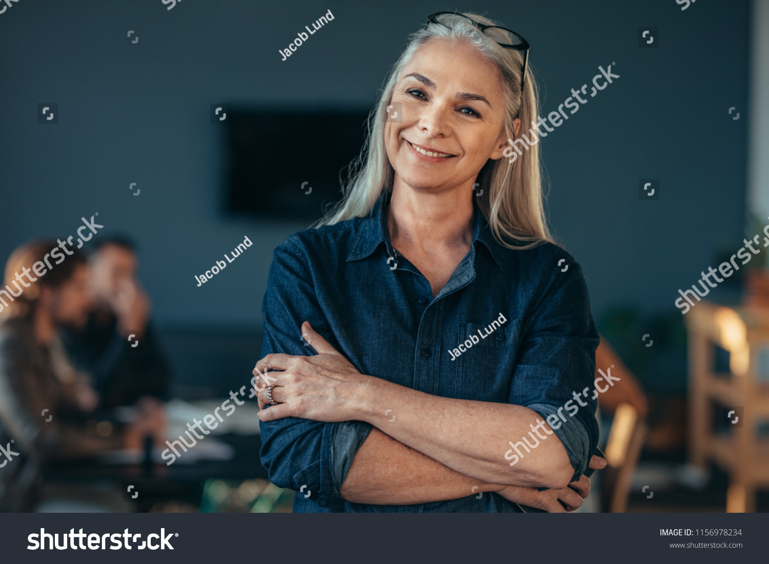 Portrait of confident senior business woman standing in office with her arms crossed. Mature female in office with colleagues discussing work at the back. #1156978234