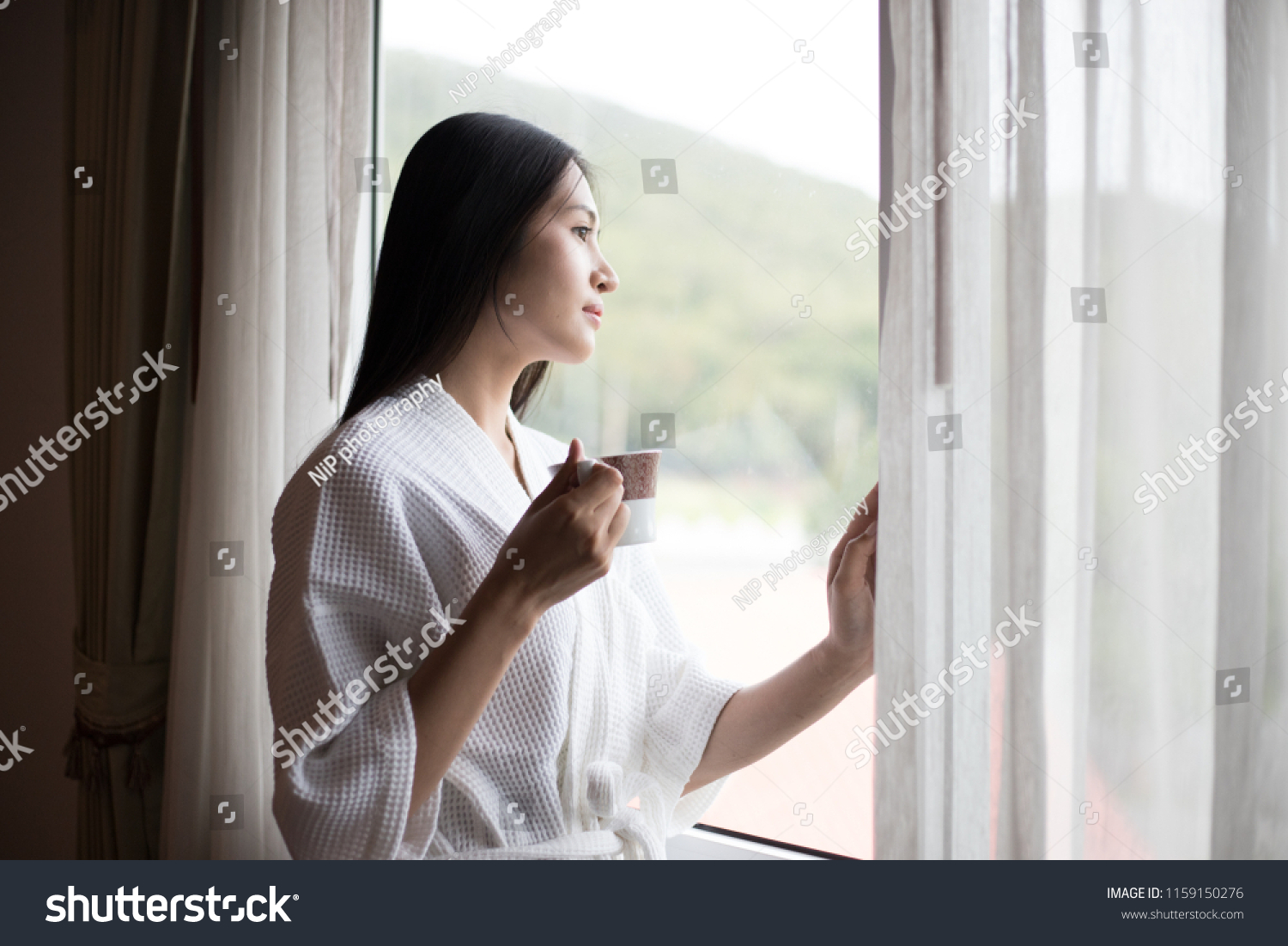 wake up :Woman in the morning holding a cup of tea or coffee and looking at the sunrise standing near the window in her home #1159150276