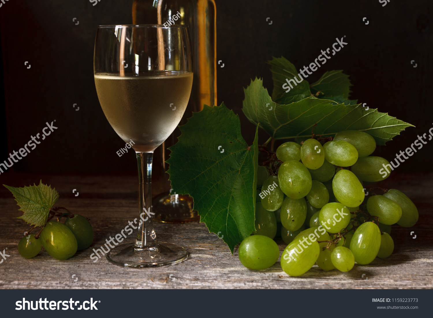 Green grapes with leaves, glass, bottle of white wine on vintage wooden background. Close-up, selective focus. Still life in vintage stile. Dark photo. #1159223773
