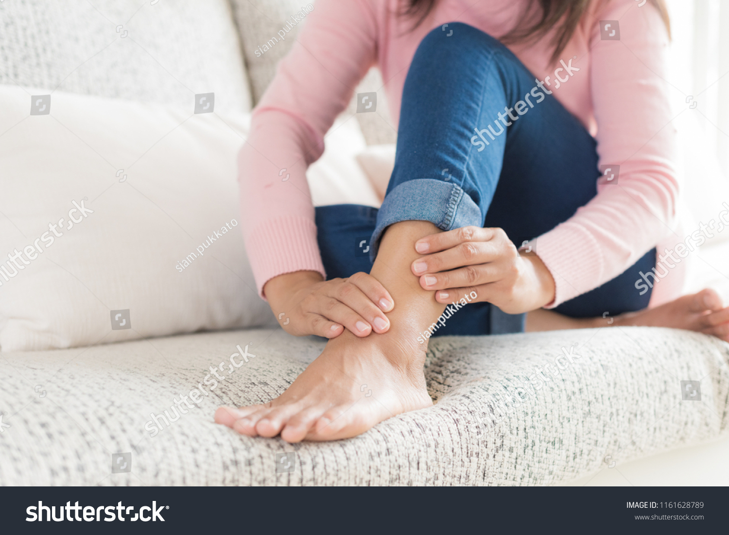 Closeup woman sitting on sofa holds her ankle injury, feeling pain. Health care and medical concept. #1161628789
