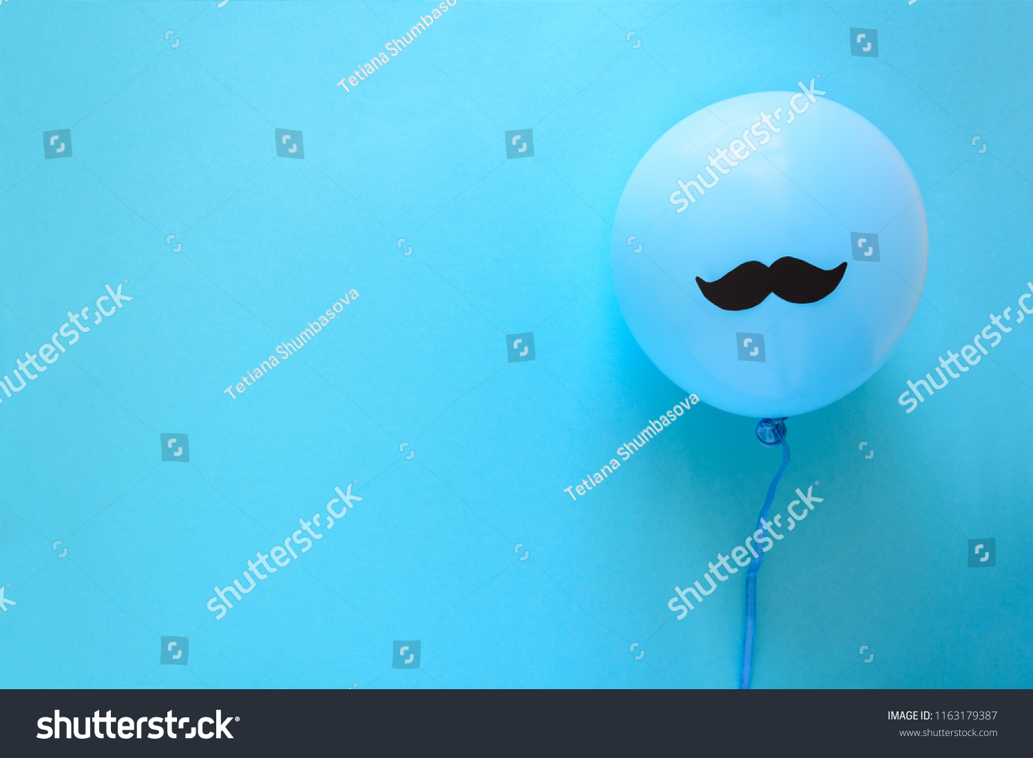 Hand holding blue balloon with a paper mustache on blue paper background. Cut out style. Father's day or mustache day concept. Top view. Flat lay. Copy space #1163179387