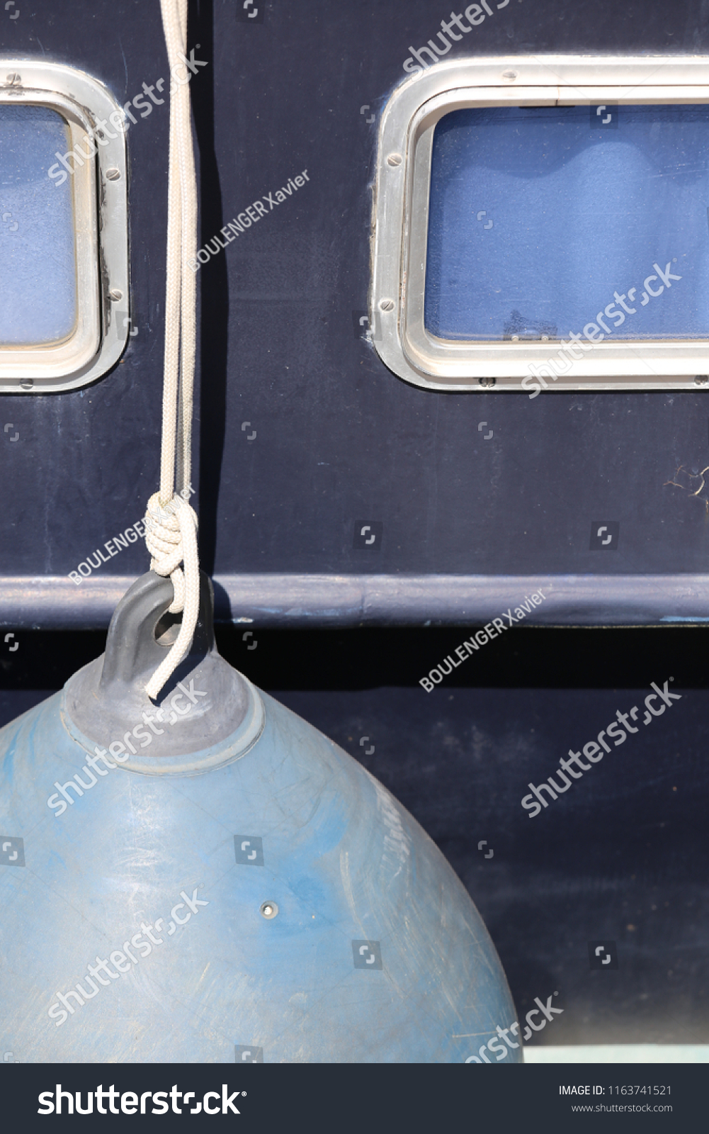 Close up outdoor view of part of a black boat hull with two white glass windows, blue curtains, a rope with a rounding float. Abstract image of maritime equipment, Bright surface and round shapes.  #1163741521