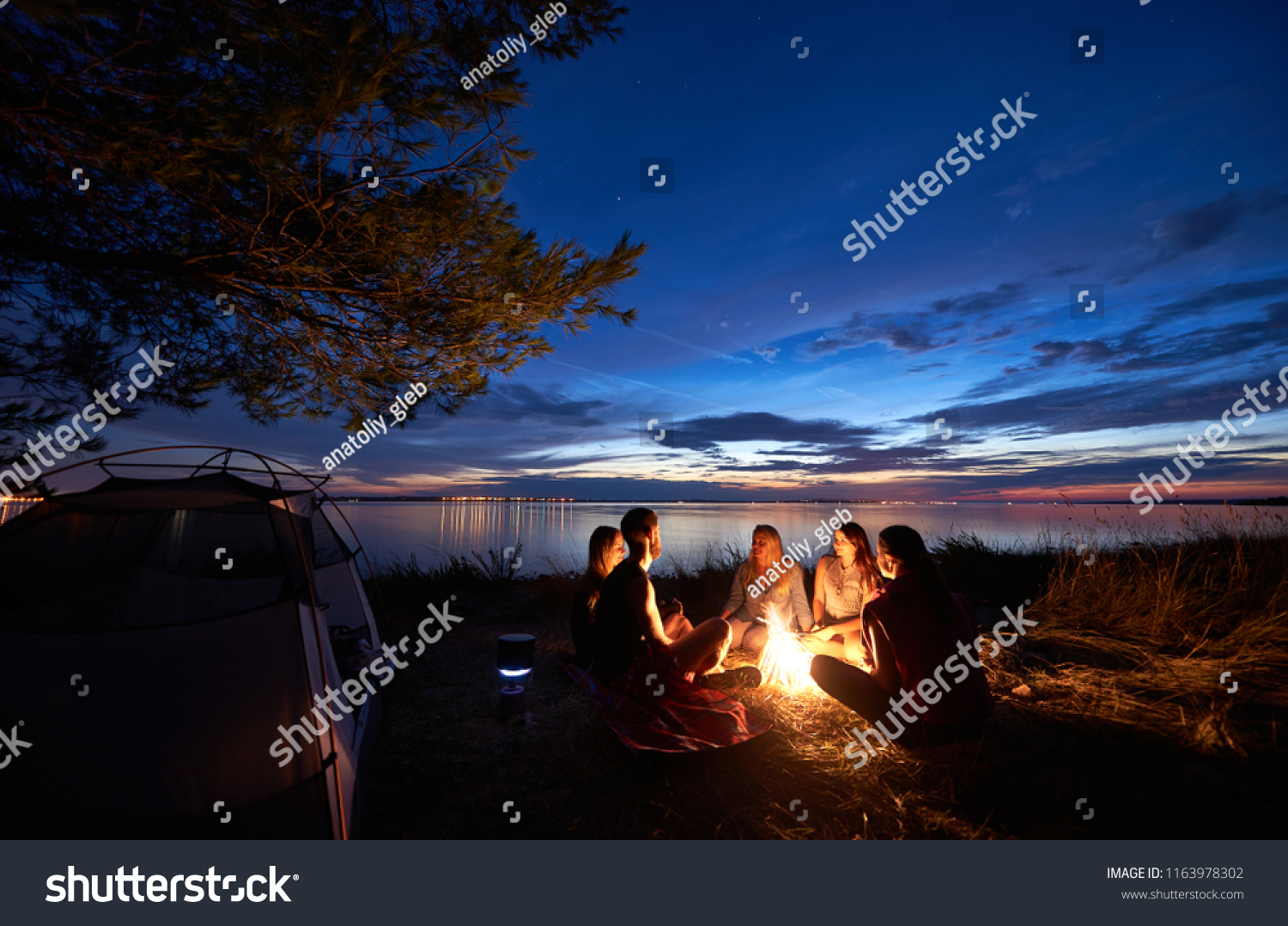 Night summer camping on sea shore. Group of five young tourists sitting on the beach around campfire near tent under beautiful blue evening sky. Tourism, friendship and beauty of nature concept. #1163978302