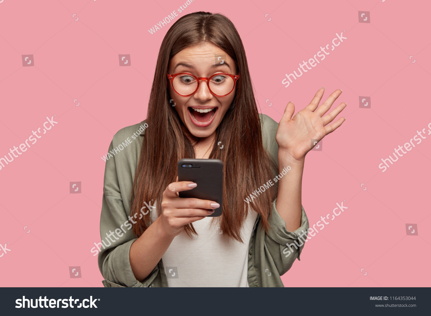 Overjoyed happy excited female glad to recieve text message informing about salary, rejoices good news, stares at mobile phone, gestures actively from happiness, stands indoor alone. Reaction concept #1164353044