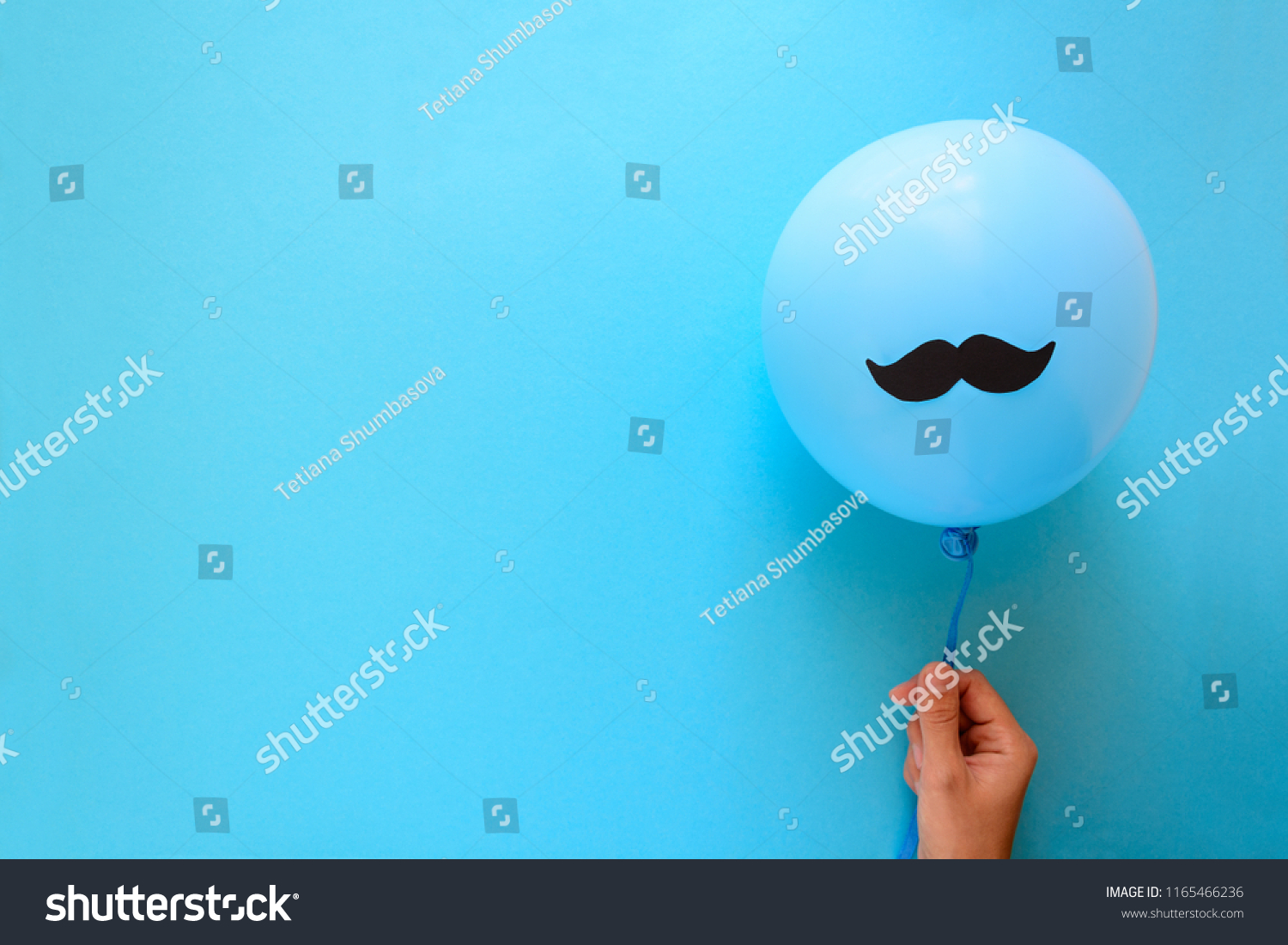 Hand holding blue balloon with a paper mustache on blue paper background. Cut out style. Father's day or man health concept. Top view. Copy space #1165466236