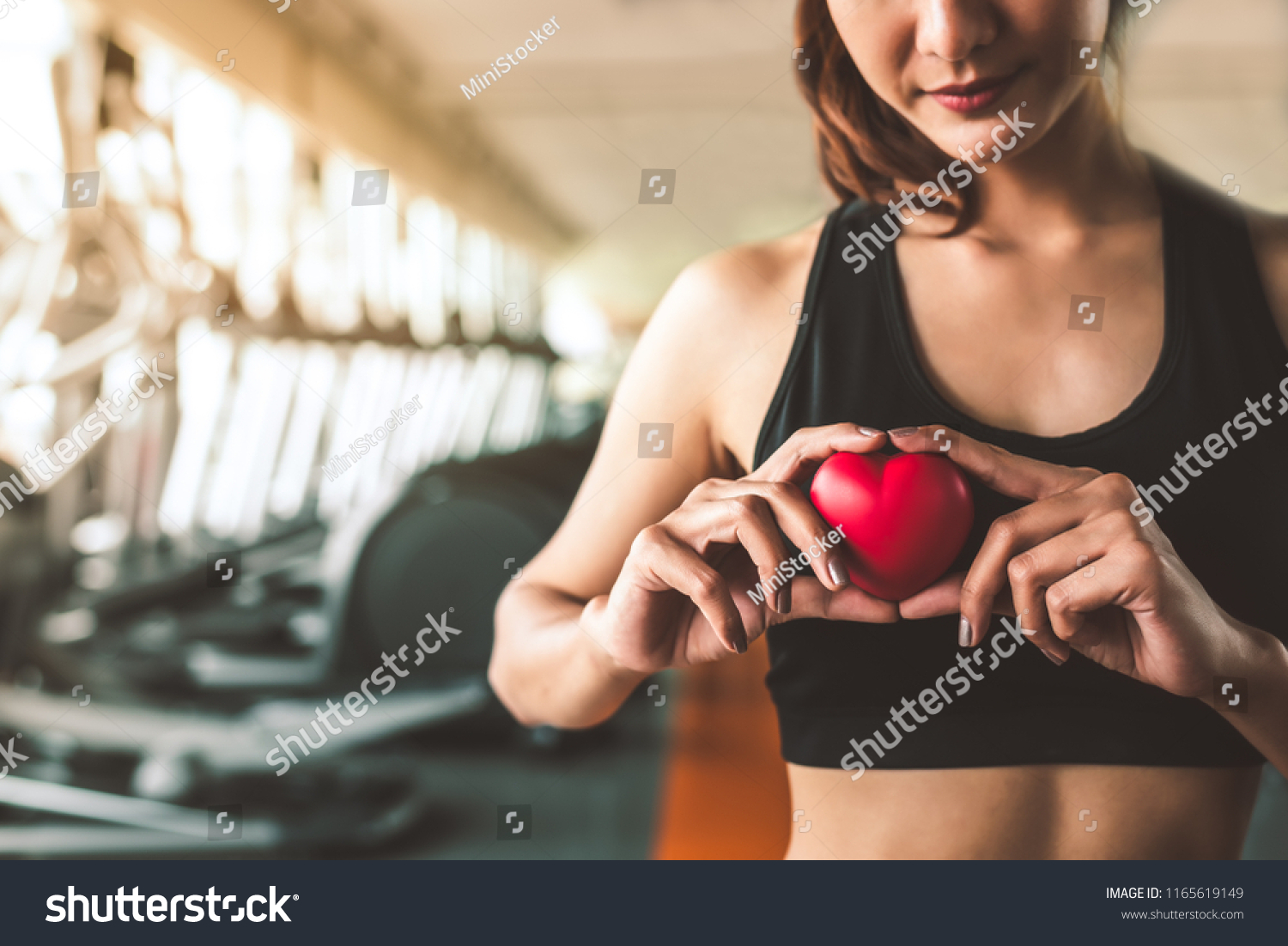 Happy sport woman holding red heart in fitness gym club. Medical cadio heart strength training lifestyle. Pretty female sport girl workout exercise. Cardiac healthy and wellbeing. Massage ball in hand #1165619149