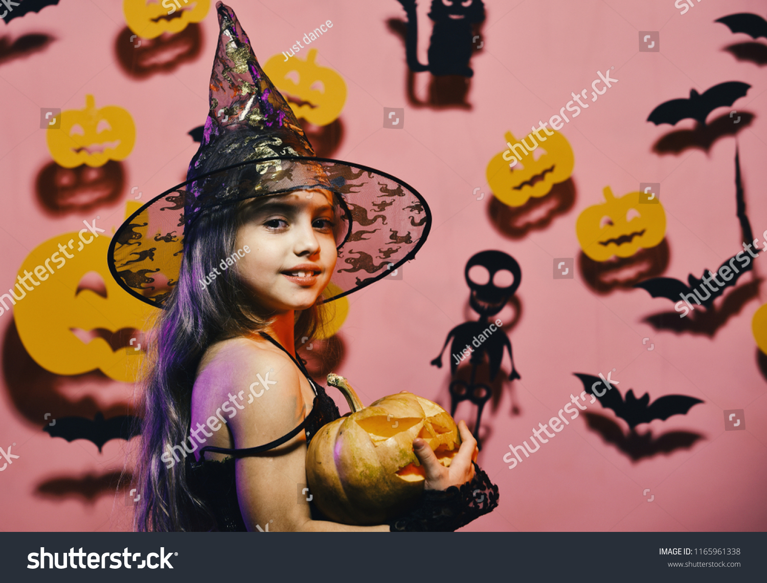 Little witch wearing black hat. Halloween party and decorations concept. Girl with smiling face on pink background with bats and pumpkins decor. Kid in spooky witches costume holds carved pumpkin #1165961338