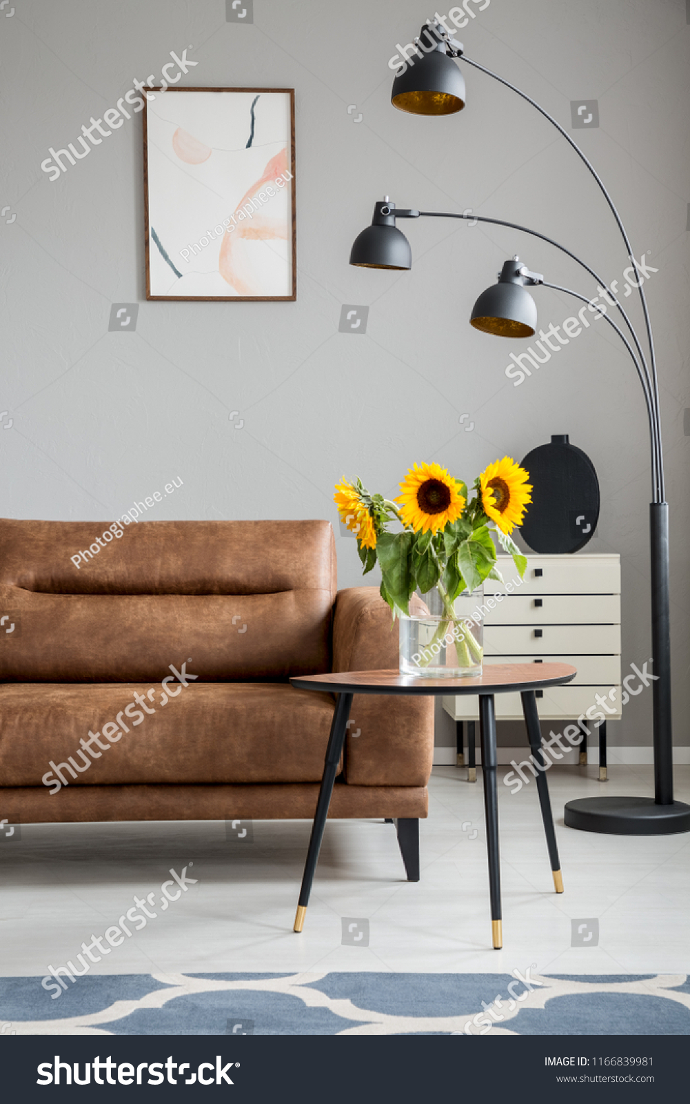 Sunflowers on wooden table next to brown sofa and black lamp in flat interior with poster. Real photo #1166839981