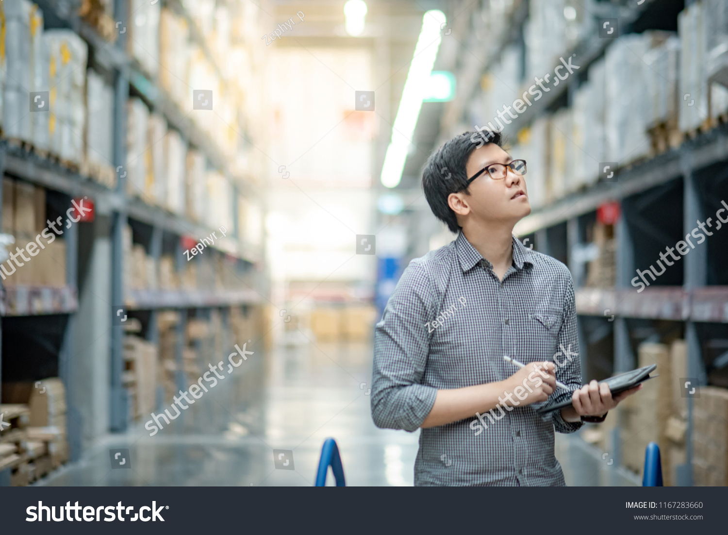 Asian manager man doing stocktaking of products in cardboard box on shelves in warehouse using digital tablet and pen. Male professional assistant checking stock in factory. Physical inventory count. #1167283660