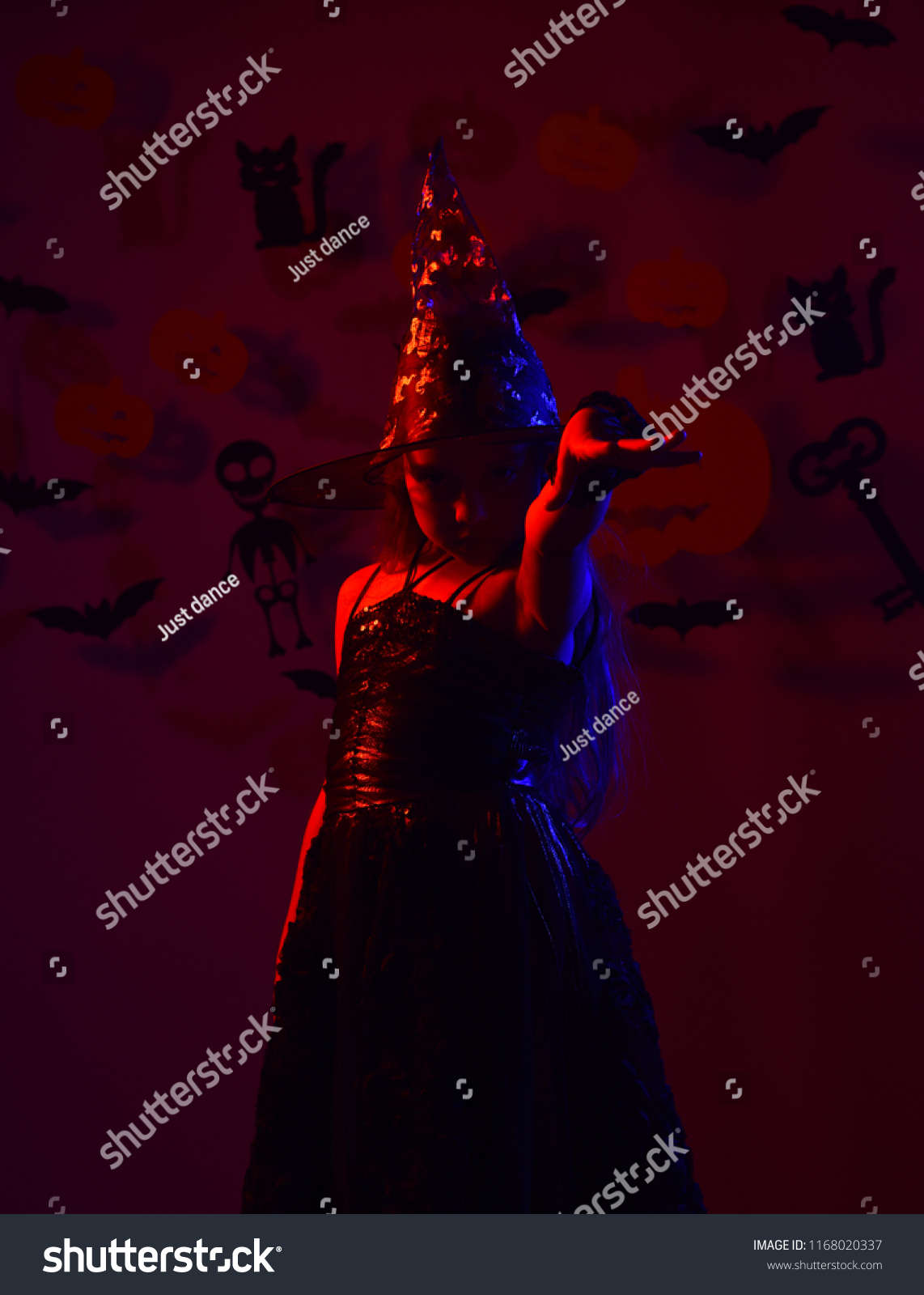 Little witch wearing black hat. Halloween party and decorations concept. Girl with confident face on bloody red background with decor. Kid in spooky witches costume poses in red light #1168020337