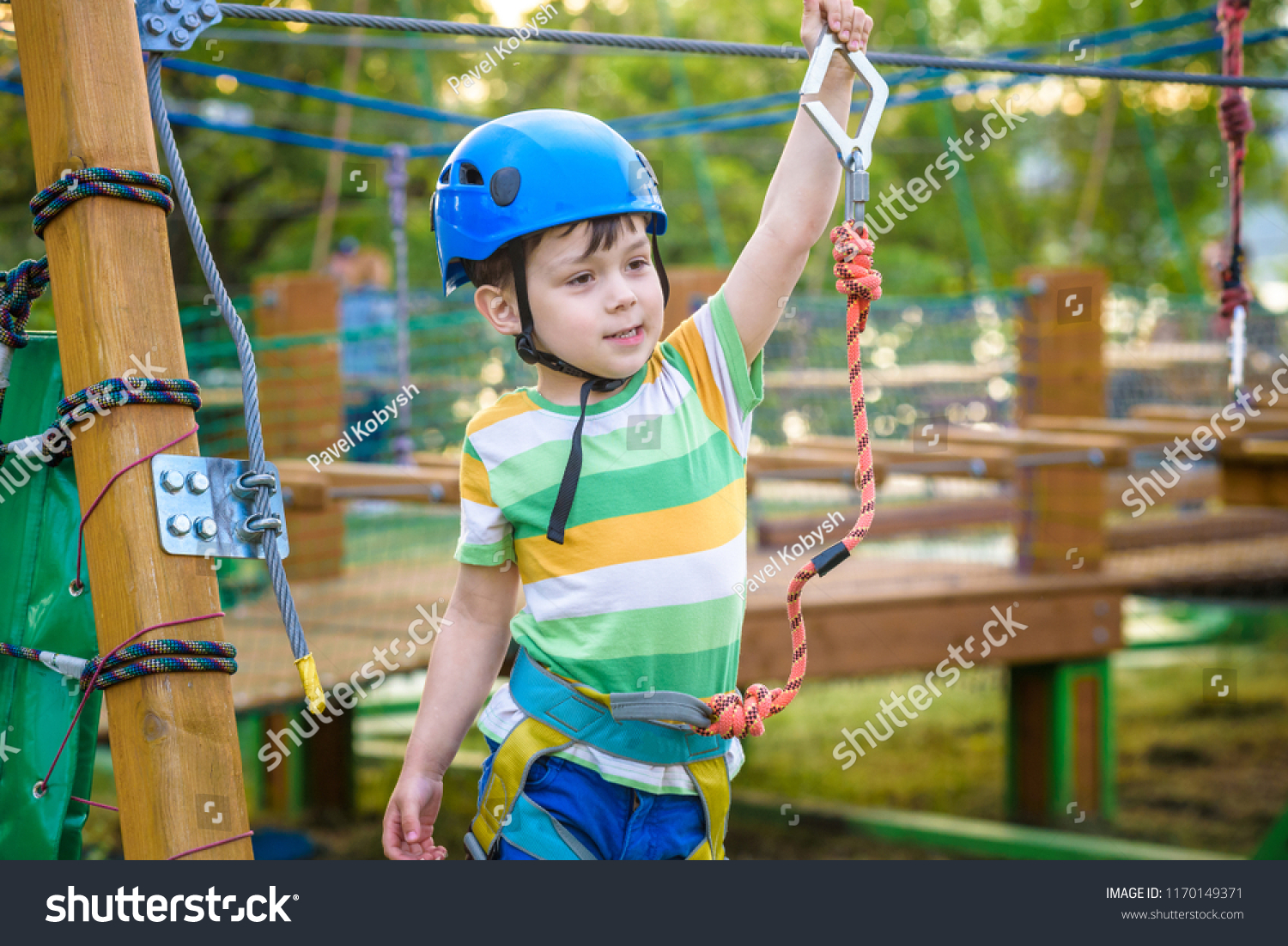 Little cute boy enjoying activity in a climbing adventure park on a summer sunny day. toddler climbing in a rope playground structure. Safe Climbing extreme sport with helmet and Carabiner. insurance #1170149371
