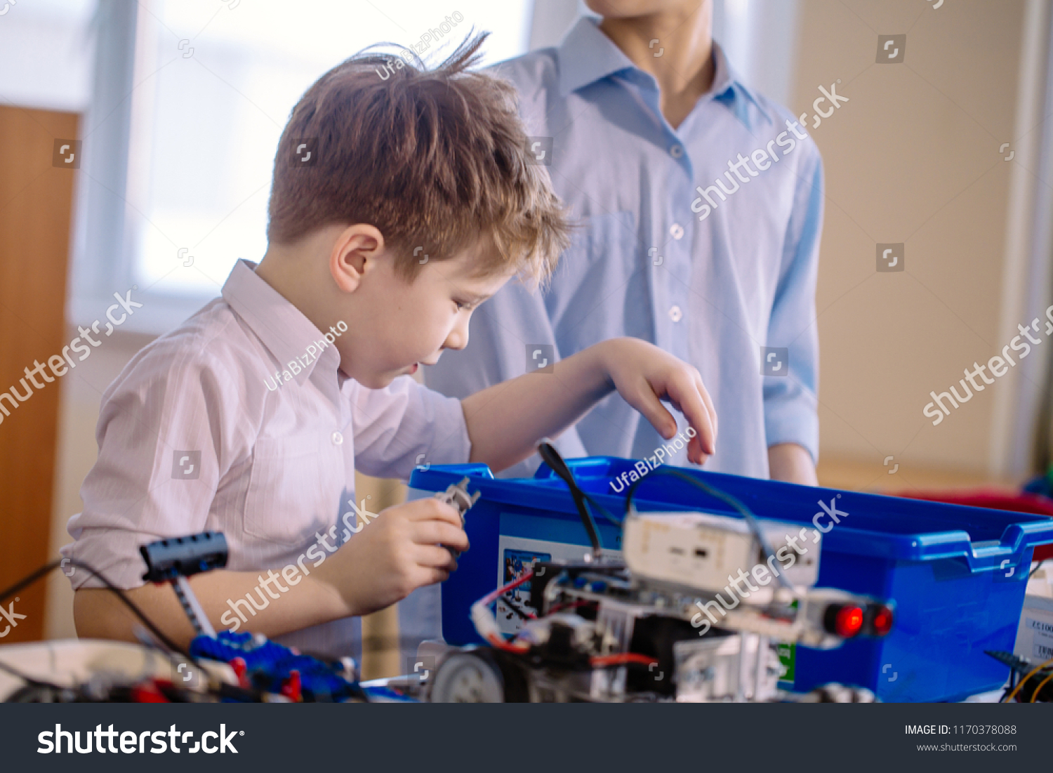 Fair haired cheerful little boy making a robot from metal parts and microcircuits, his brother helps him, close up. Happy emotion and enjoyment. #1170378088