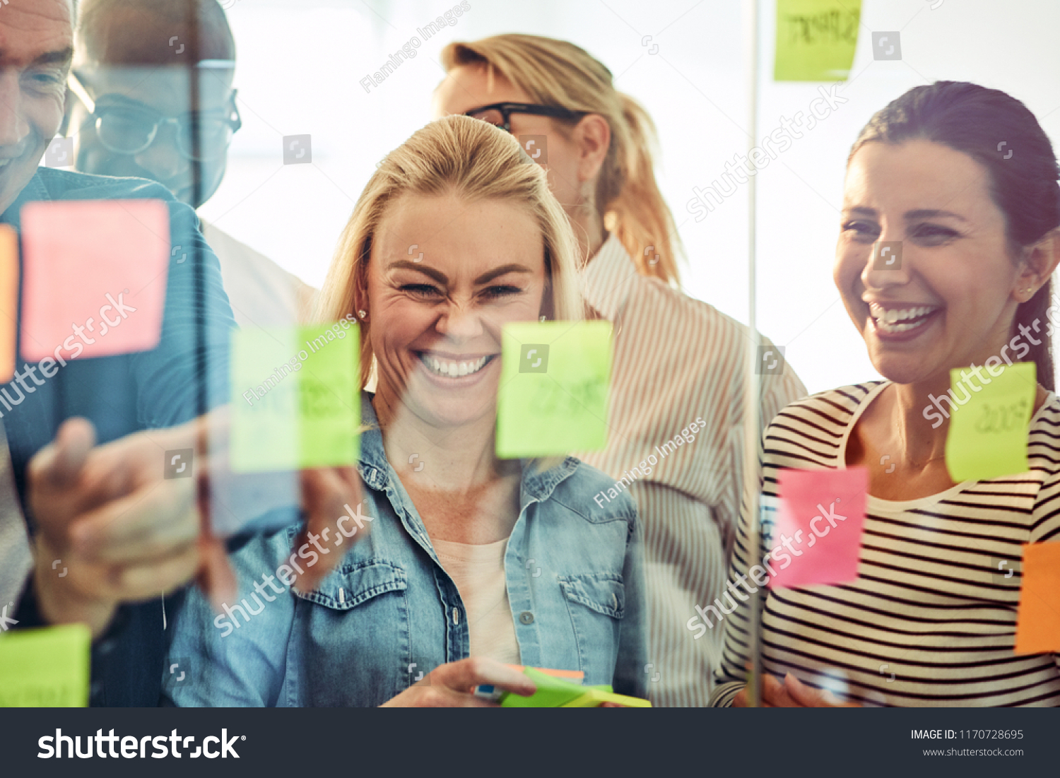Diverse group of businesspeople laughing while standing in an office brainstorming together with sticky notes on a glass wall #1170728695