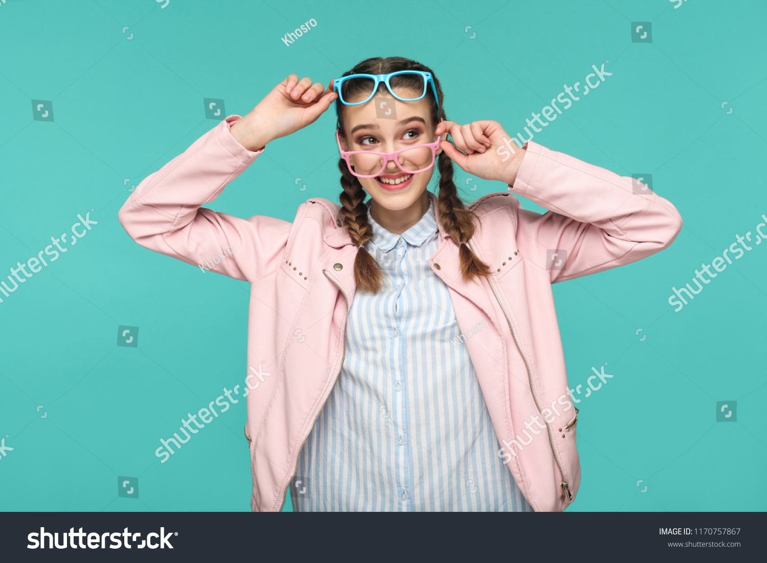 Happy funny girl in casual or hipster style, pigtail hairstyle, standing, holding two blue and pink glasses and looking away with toothy smile, Indoor studio shot, isolated on blue or green background #1170757867