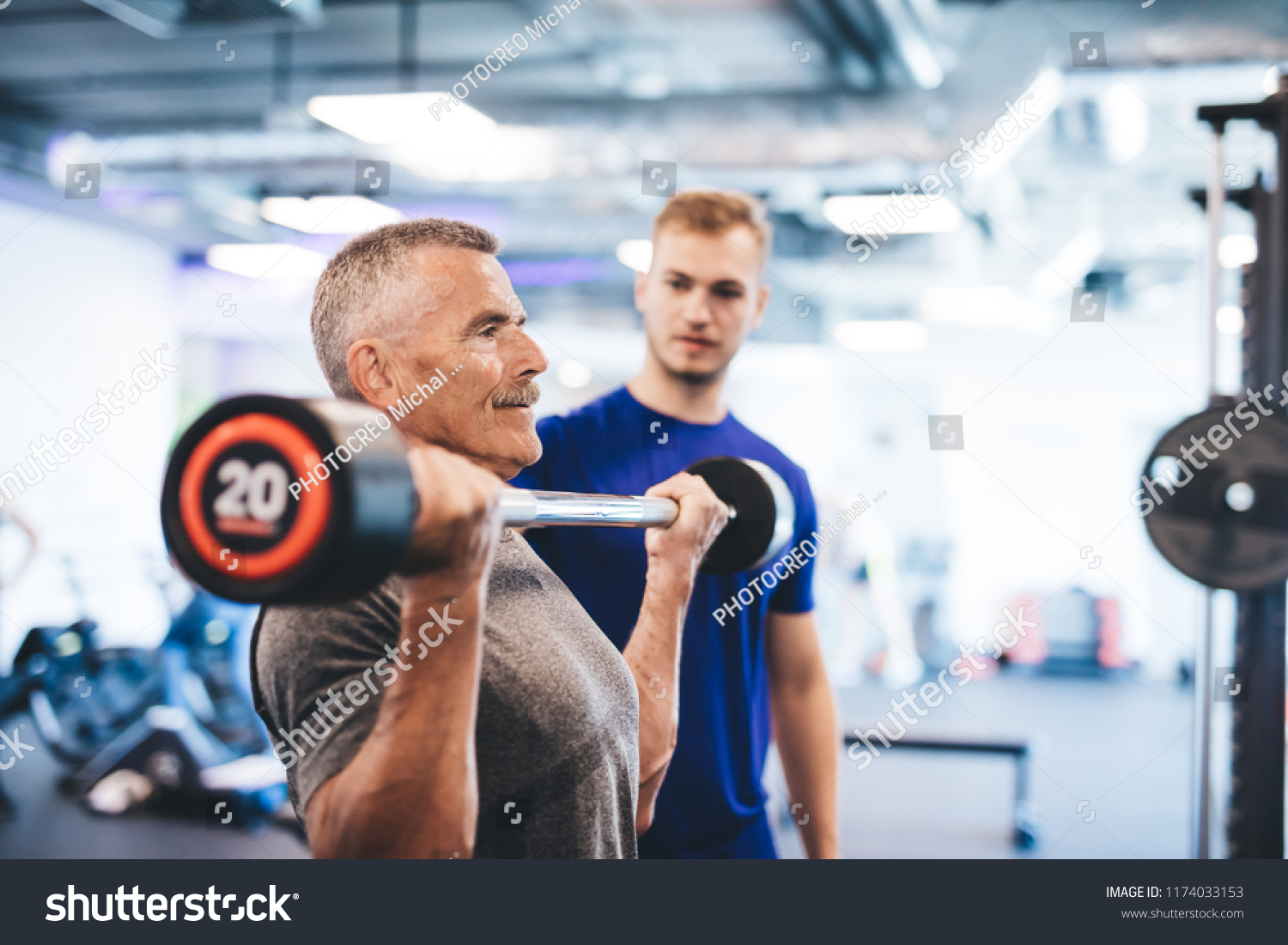 Older man lifting weights, supervised by gym assistant. Sporty lifestyle of elderly people. #1174033153