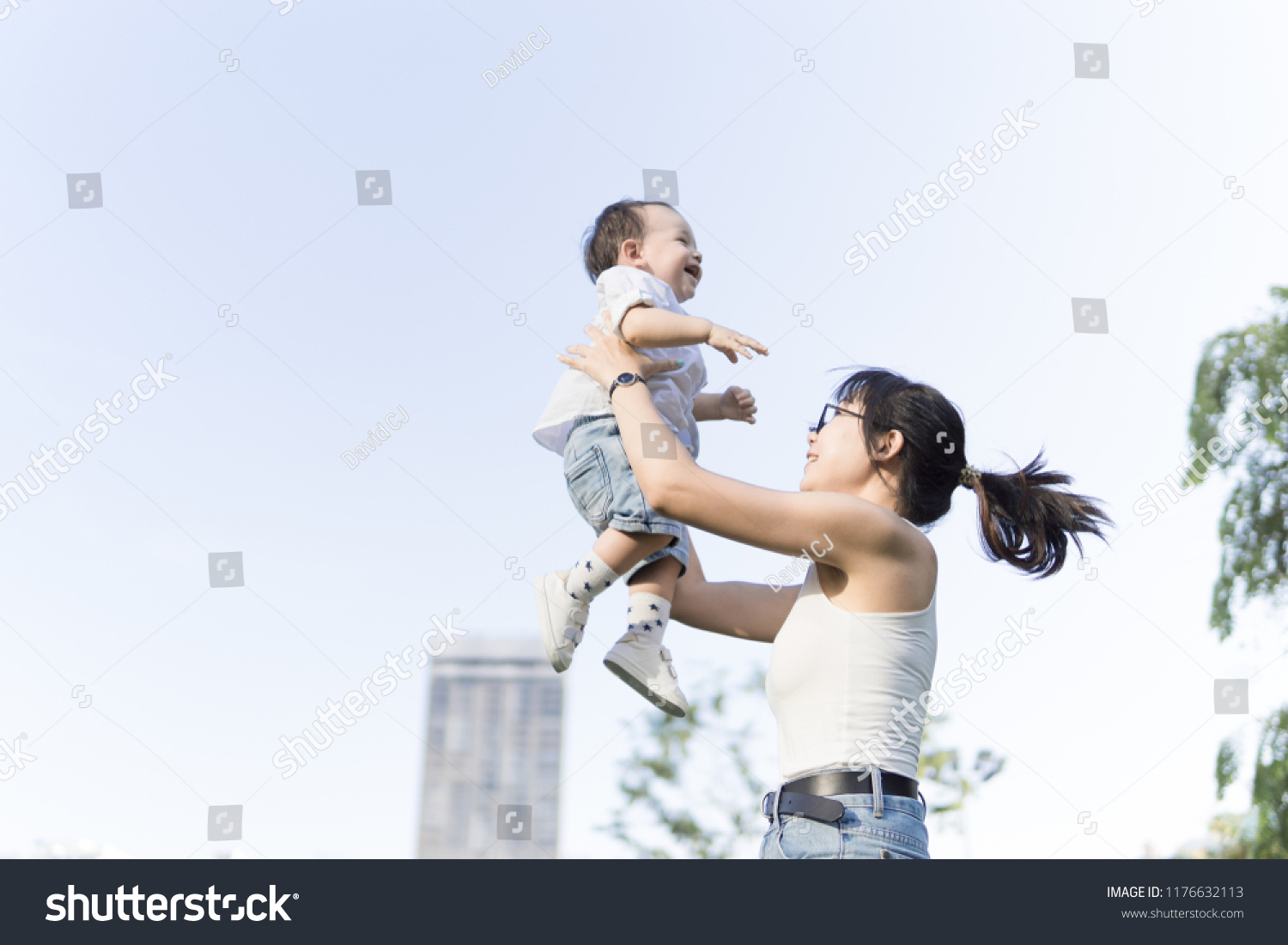 Young Asian mother holding and playing launching his happy baby boy in the air while he similes and laughs outdoors #1176632113
