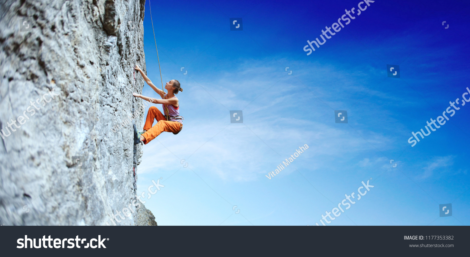 side view of young slim woman rock climber in bright orange pants climbing on the cliff. a woman climbs on a vertical rock wall on the blue sky background #1177353382