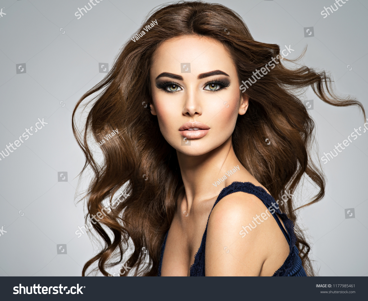 Beautiful caucasian woman with long brown curly hair. Portrait of a pretty young adult girl. Sexy face of an attractive  lady posing at studio over grey background. #1177985461