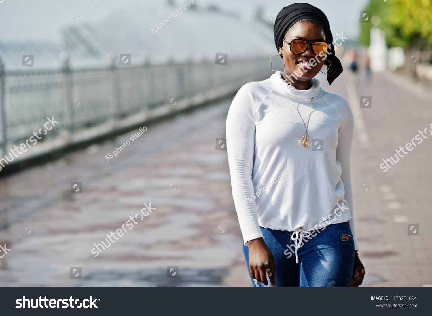 African muslim girl in sunglasses, black hijab, white sweatshirt and jeans posed outdoor against fountain. #1178271094