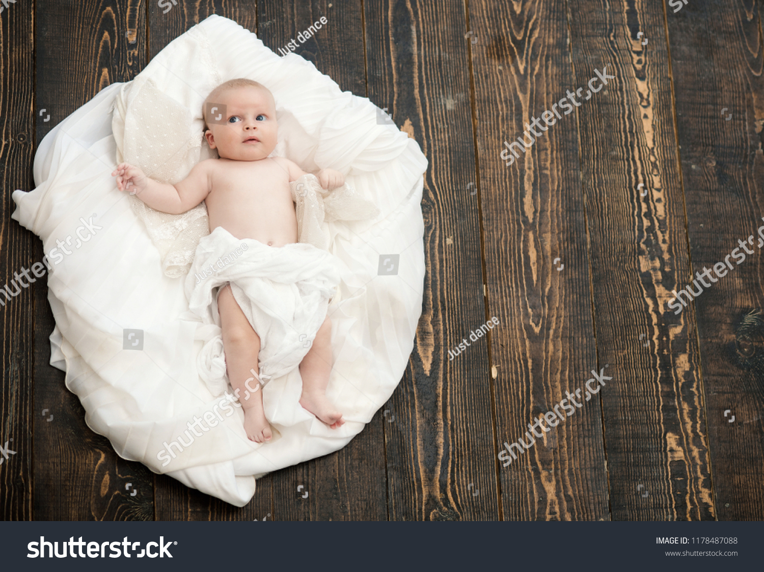 Newborn toddler with blue eyes and curious face on wooden background. Infant covered with white blanket. Baby lying on white soft duvet designed as cloud or nest. Childhood and innocence concept #1178487088