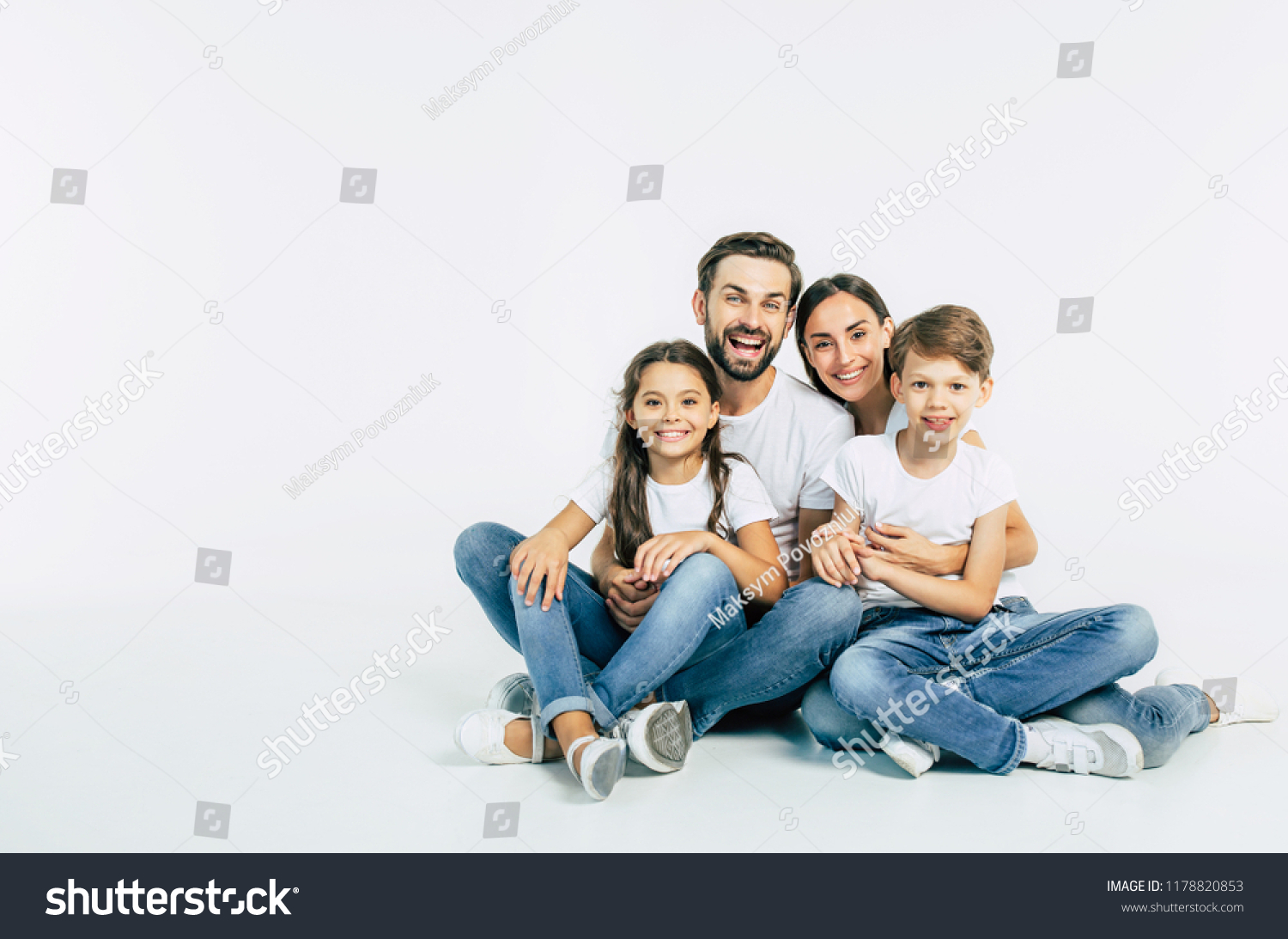 Cool team. Beautiful and happy smiling young family in white T-shirts are hugging and have a fun time together while sitting on the floor and looking on camera. #1178820853