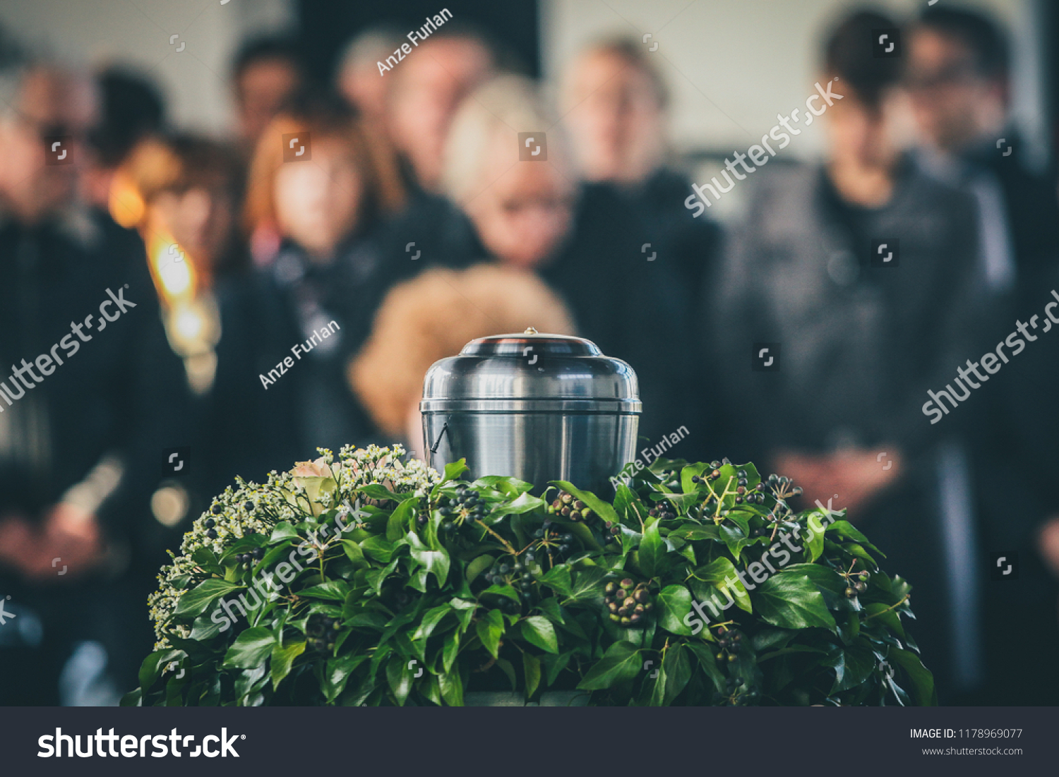 A metal urn with ashes of a dead person on a funeral, with people mourning in the background on a memorial service. Sad grieving moment at the end of a life. Last farewell to a person in an urn. #1178969077