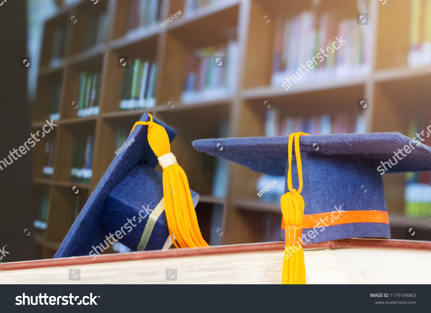 Graduate or Education knowledge learning study abroad concept : Graduation cap on opening textbook in old library stack of text literature on table with bookshelves in university college, light flare #1179104863