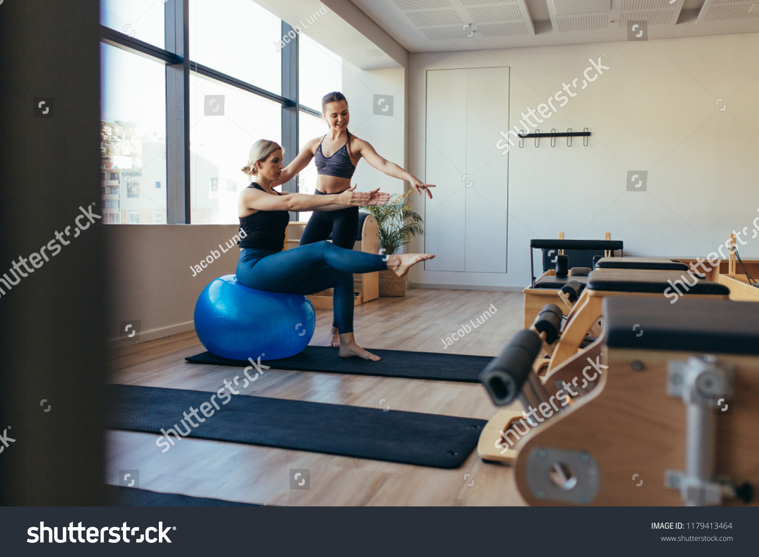 Female pilates instructor training a woman in pilates workout. Woman doing pilates workout sitting on a fitness ball in a gym. #1179413464