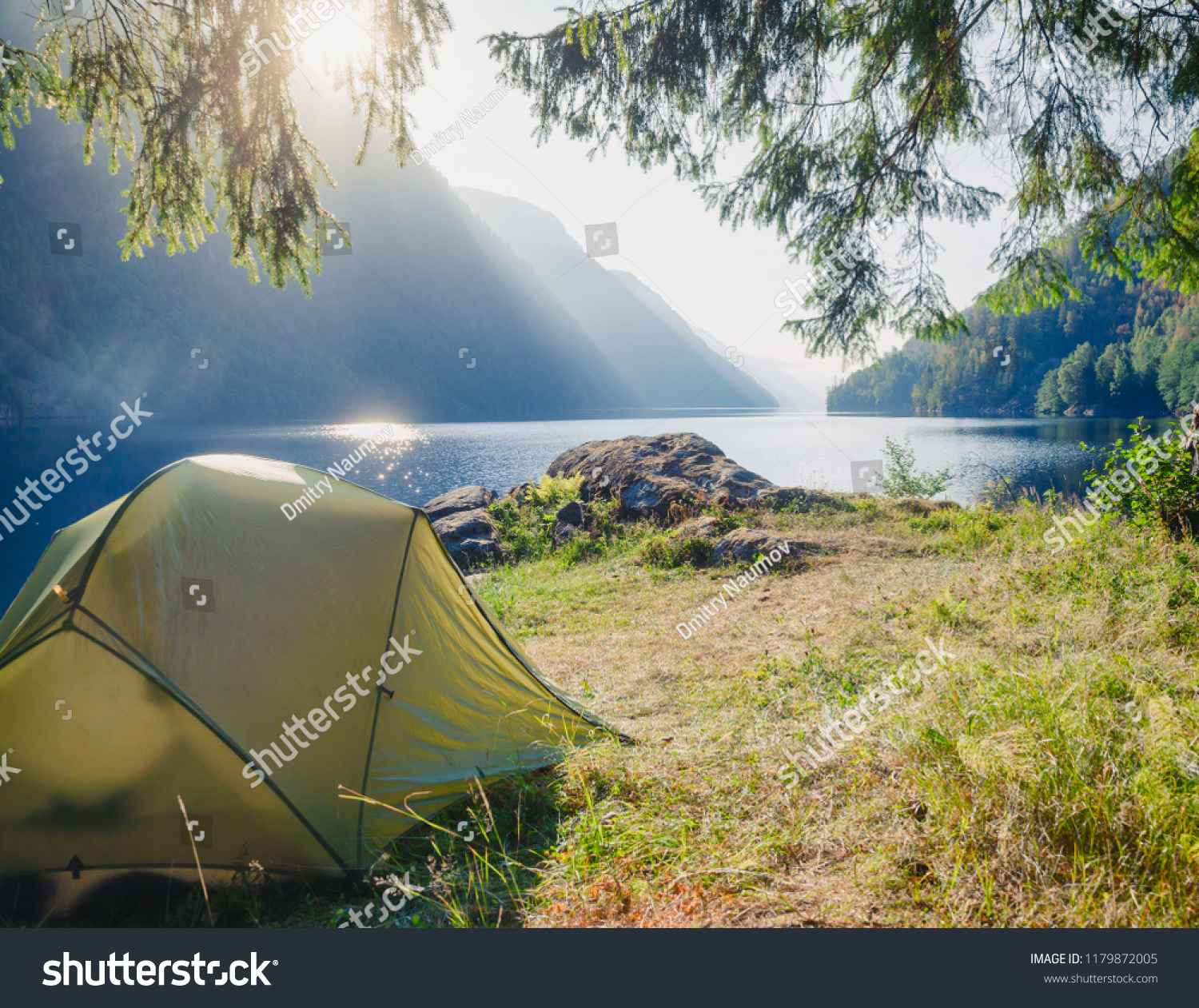 Sunlit camping tent at scenic campsite on a lake shore with mountain range in background - wild camping in Norway #1179872005