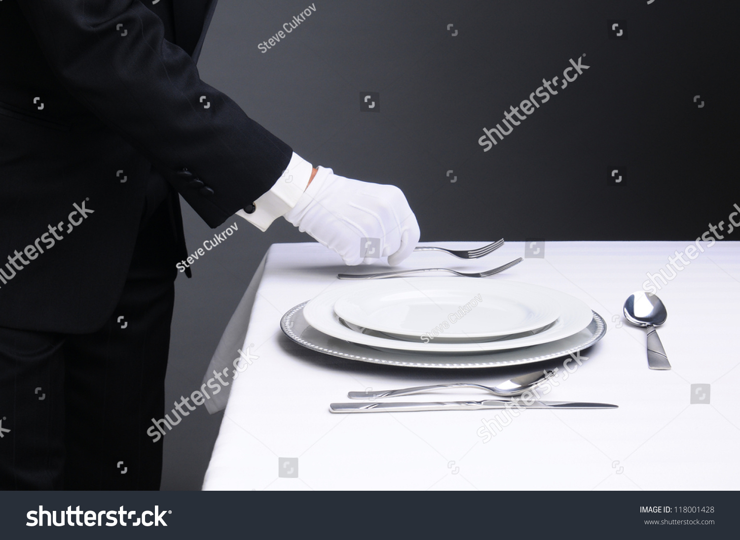 Closeup of a waiter in a tuxedo setting a formal dinner table. Horizontal format on a light to dark gray background. Man is unrecognizable. #118001428