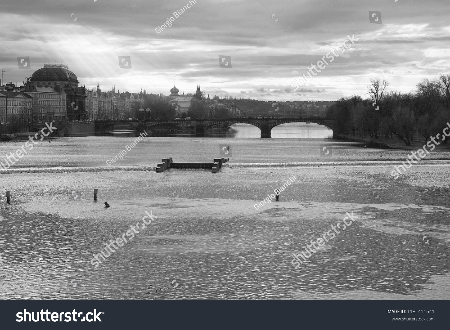 With Charles Bridge and New Town district of Prague in the background, a beautiful landscape of Vltava River in black and white with sun rays spreading through the clouds #1181411641