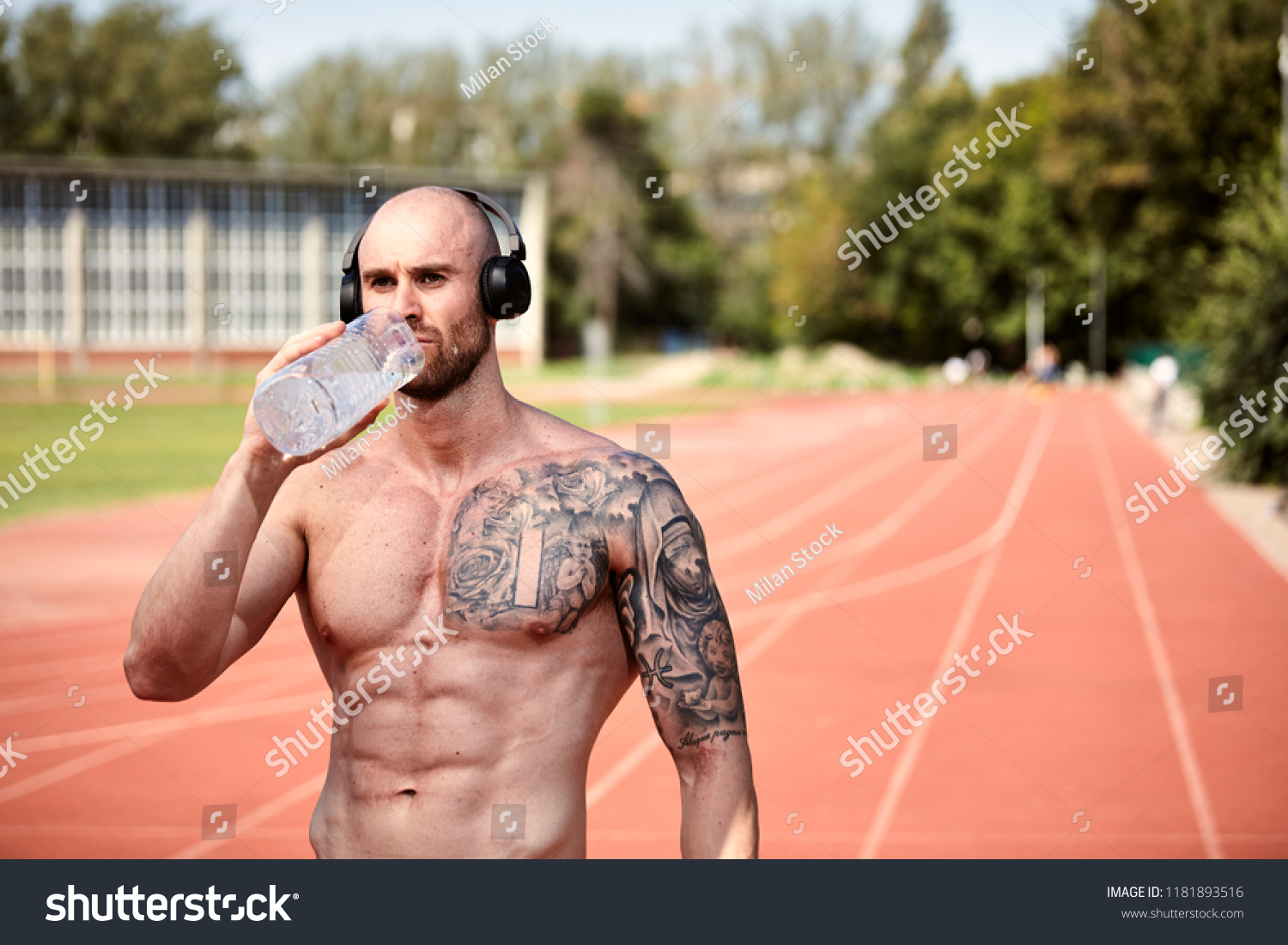 one young strong tattooed man shirtless barechestedness, bodybuilder, 30-35 years, drinking water from bottle. Outdoors, sports venue, running tracks (out of focus). upper body shot. #1181893516