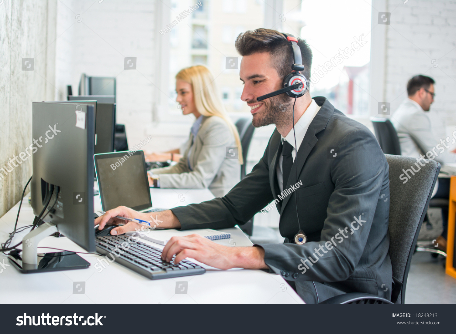 Portrait of a smiling young handsome man in formalwear with headset using computer in a office #1182482131