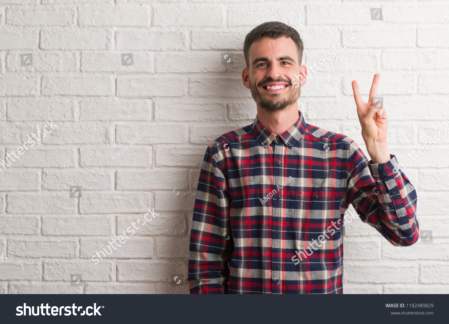 Young adult man standing over white brick wall showing and pointing up with fingers number two while smiling confident and happy. #1182489829