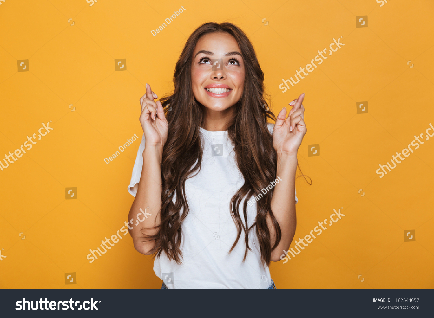 Portrait of a smiling young girl with long brunette hair standing over yellow background, holding fingers crossed for good luck #1182544057