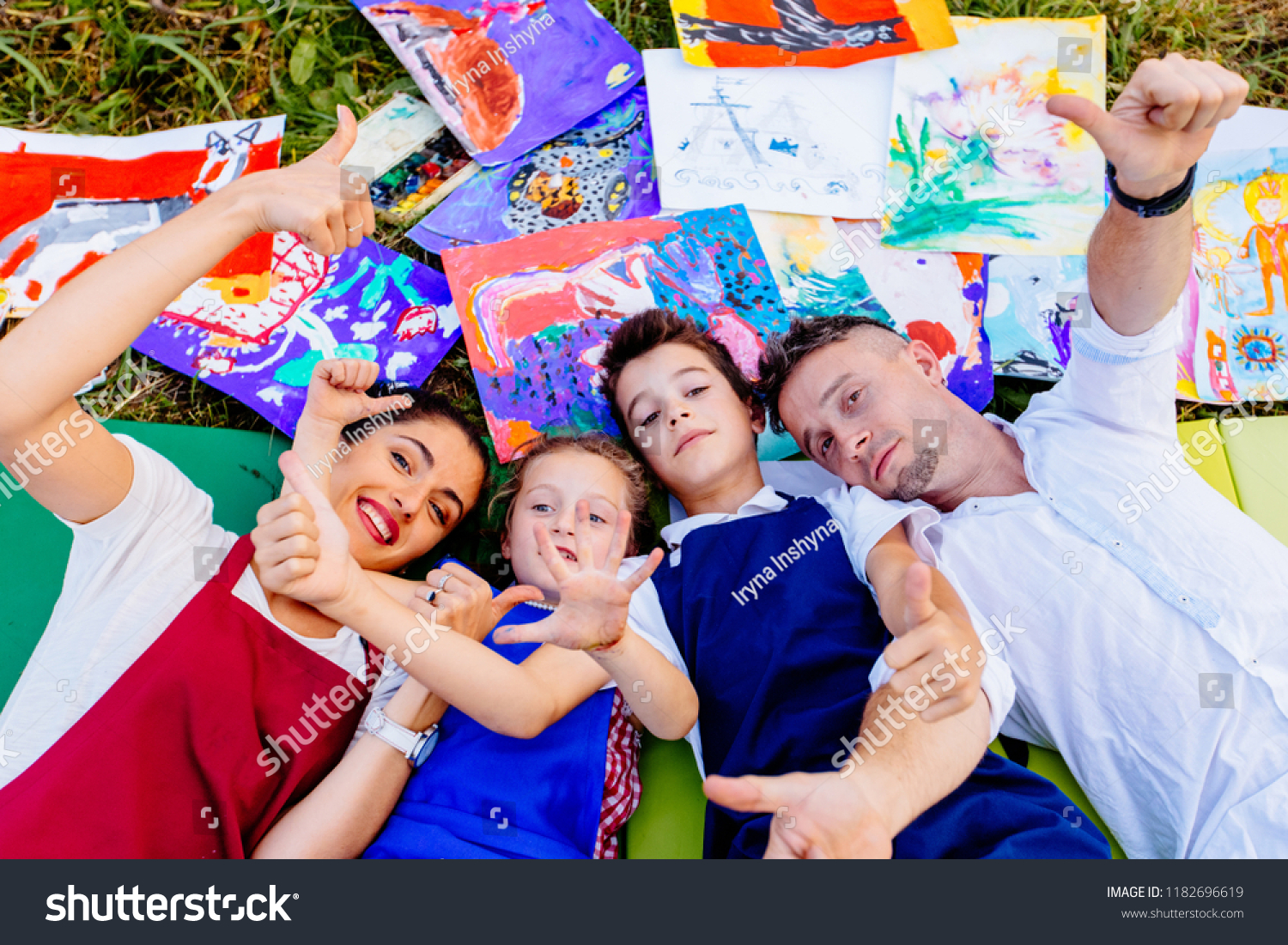 Smiling creative family paitners of mother, father, and two children in aprons lying down on grass with drawings outside having fun with thumbs up outstrectched to the camera. #1182696619